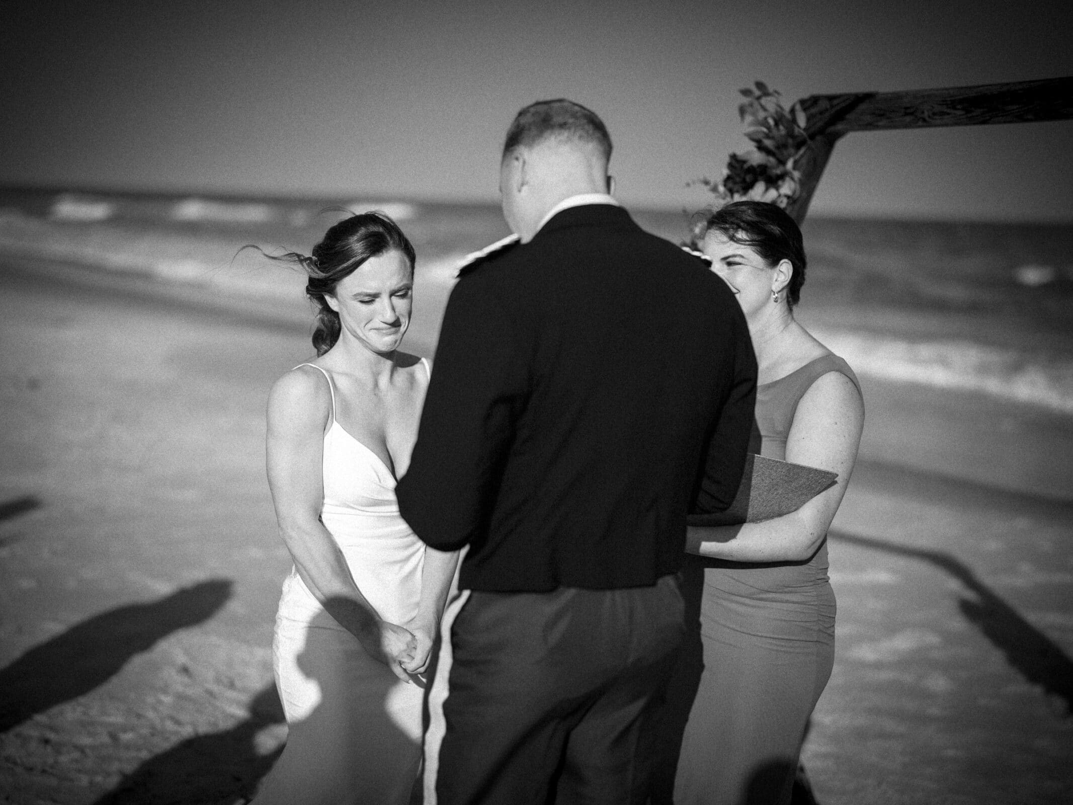 A bride and groom stand facing each other on a beach during their wedding ceremony, with an officiant beside them. The bride looks emotional, and the groom stands with his back to the camera. The ocean is in the background.