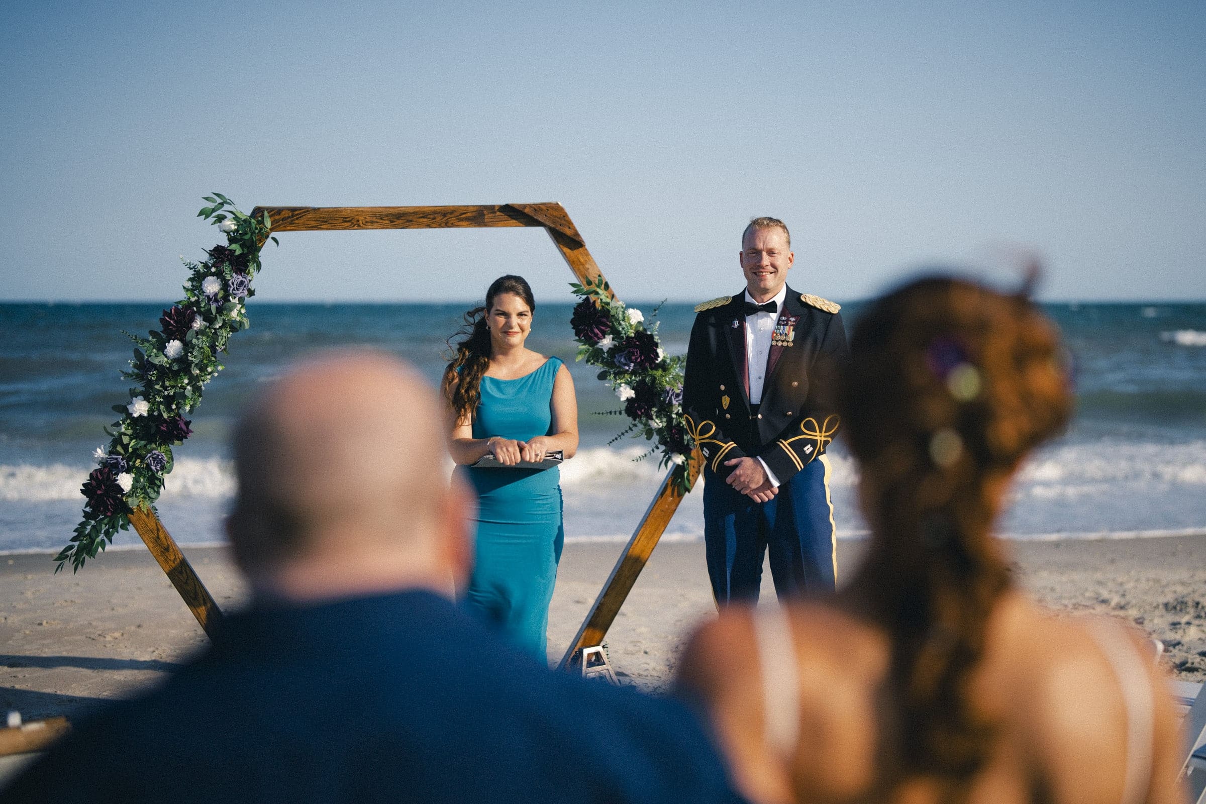 A woman in a teal dress and a man in a formal military uniform stand at a decorated wooden arch on Topsail Beach, facing two seated guests with the ocean in the background—capturing a perfect Topsail Beach elopement moment.
