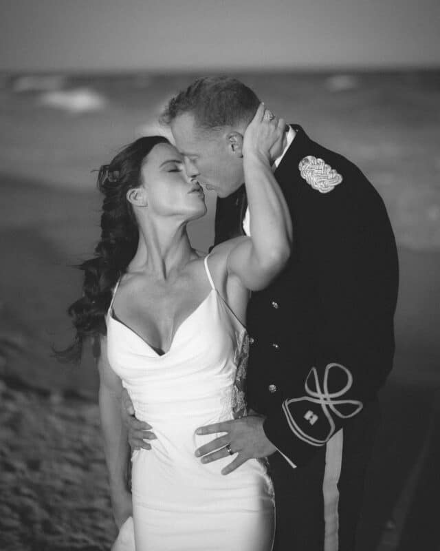 A couple shares a passionate kiss on the beach; the woman wears a white dress, and the man is in a decorated military uniform. The scene is intimate and romantic, with the ocean blurred in the background.