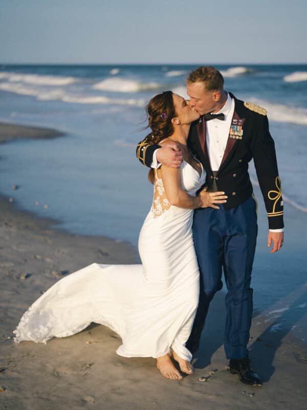 A bride in a white dress and a groom in a military uniform kiss while standing barefoot on a sandy beach with waves in the background.