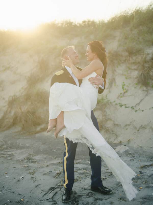 A groom in a military uniform joyfully lifts and holds his bride on a sandy beach at sunset, both smiling and gazing at each other with grass-covered dunes in the background.
