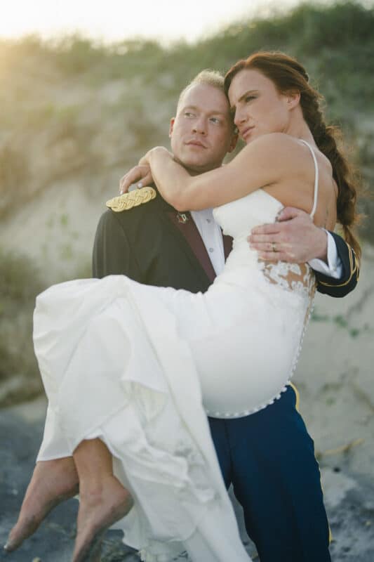A groom in formal military attire carries a bride in a white wedding dress outdoors, with sunlight shining and a sandy, grassy background. Both look into the distance with serious expressions.