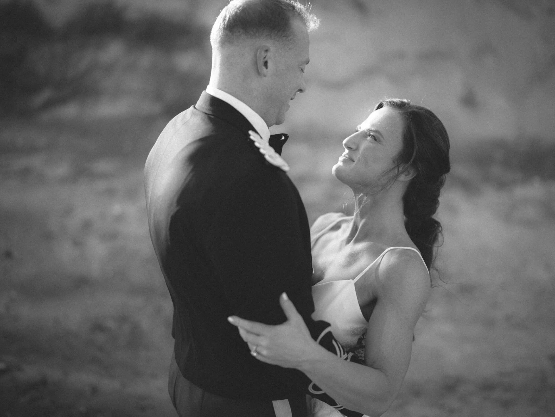 A couple dressed formally embrace and gaze into each others eyes, smiling softly. The photo is in black and white, creating a timeless, romantic atmosphere.