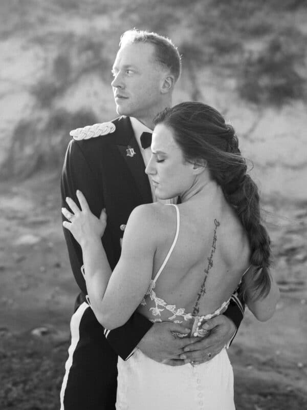 A man in a formal military uniform embraces a woman in a backless wedding dress on a beach. She has a long tattoo along her spine and both look thoughtful, gazing in different directions.