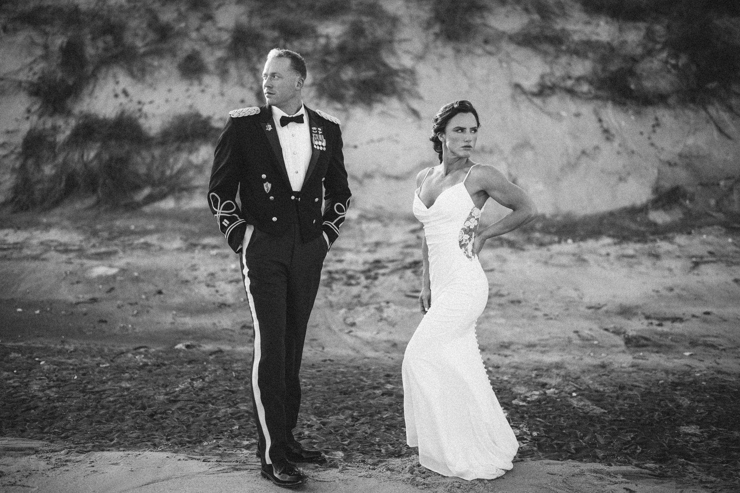 A couple stands on a sandy beach; the man wears a formal military uniform and the woman wears a white, sleeveless gown. Both pose confidently against a backdrop of dunes. The image is black and white.