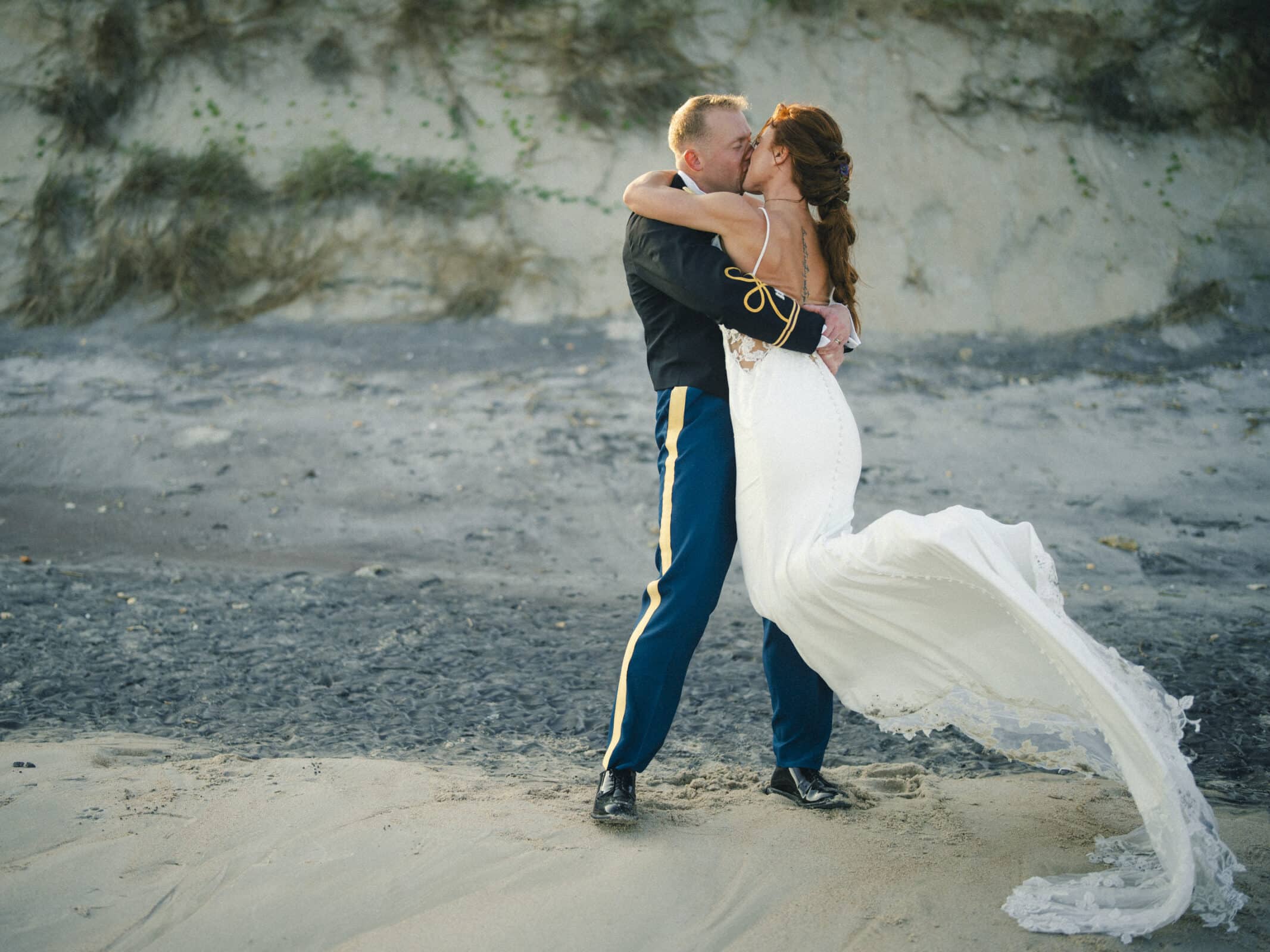A bride in a white gown and a groom in dress uniform passionately kiss and embrace on a sandy beach, with dunes and greenery in the background.