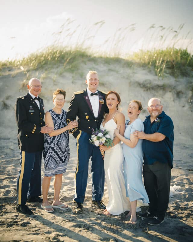 Six people stand smiling on a sandy beach at a Topsail Beach Elopement, including a couple in wedding attire, two men in military uniforms, and two others in casual and semi-formal clothing, with grasses and blue sky in the background.