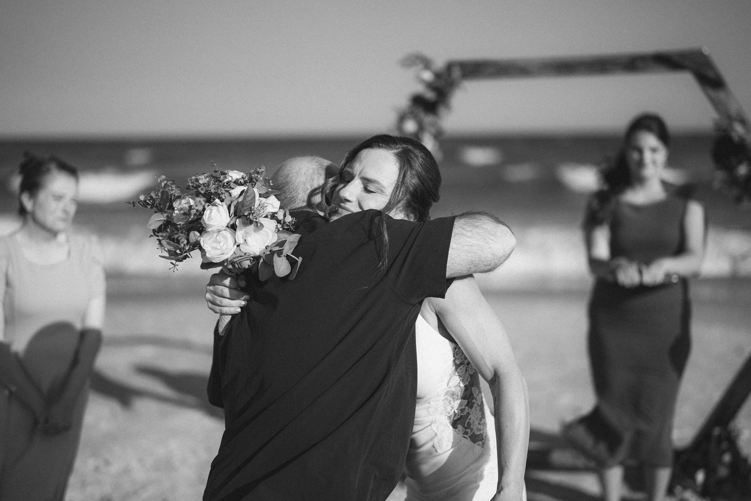 A bride hugs an older man on a beach during a wedding ceremony, holding a bouquet of flowers. Two women stand in the background, with ocean waves and a decorated wooden arch visible. The scene is in black and white.