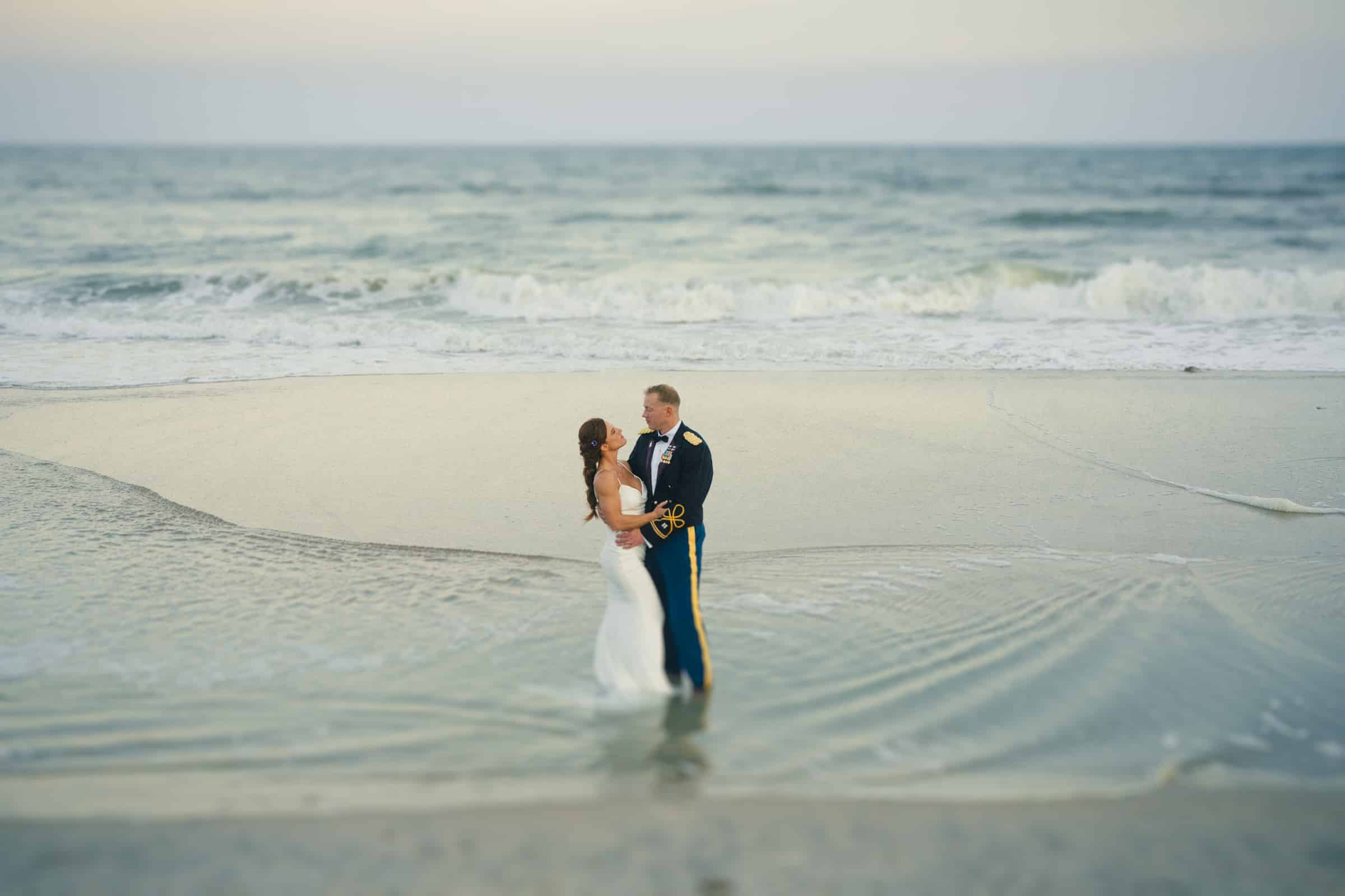A bride in a white dress and a groom in a military uniform embrace while standing barefoot in shallow water on a beach, with gentle waves and the ocean in the background.