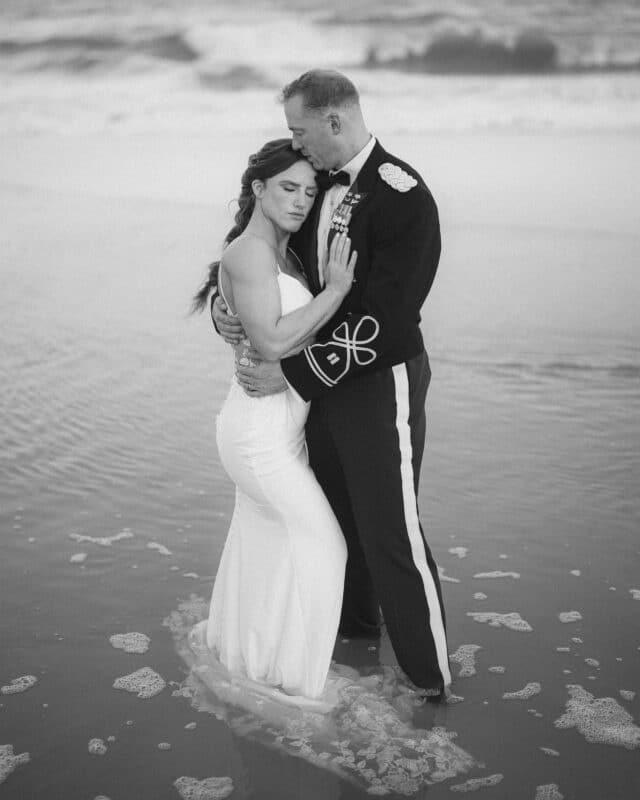 A bride in a sleeveless white gown embraces a groom in a military dress uniform as they stand ankle-deep in the ocean, eyes closed, sharing an intimate moment. The black and white photo adds a timeless, romantic feel.