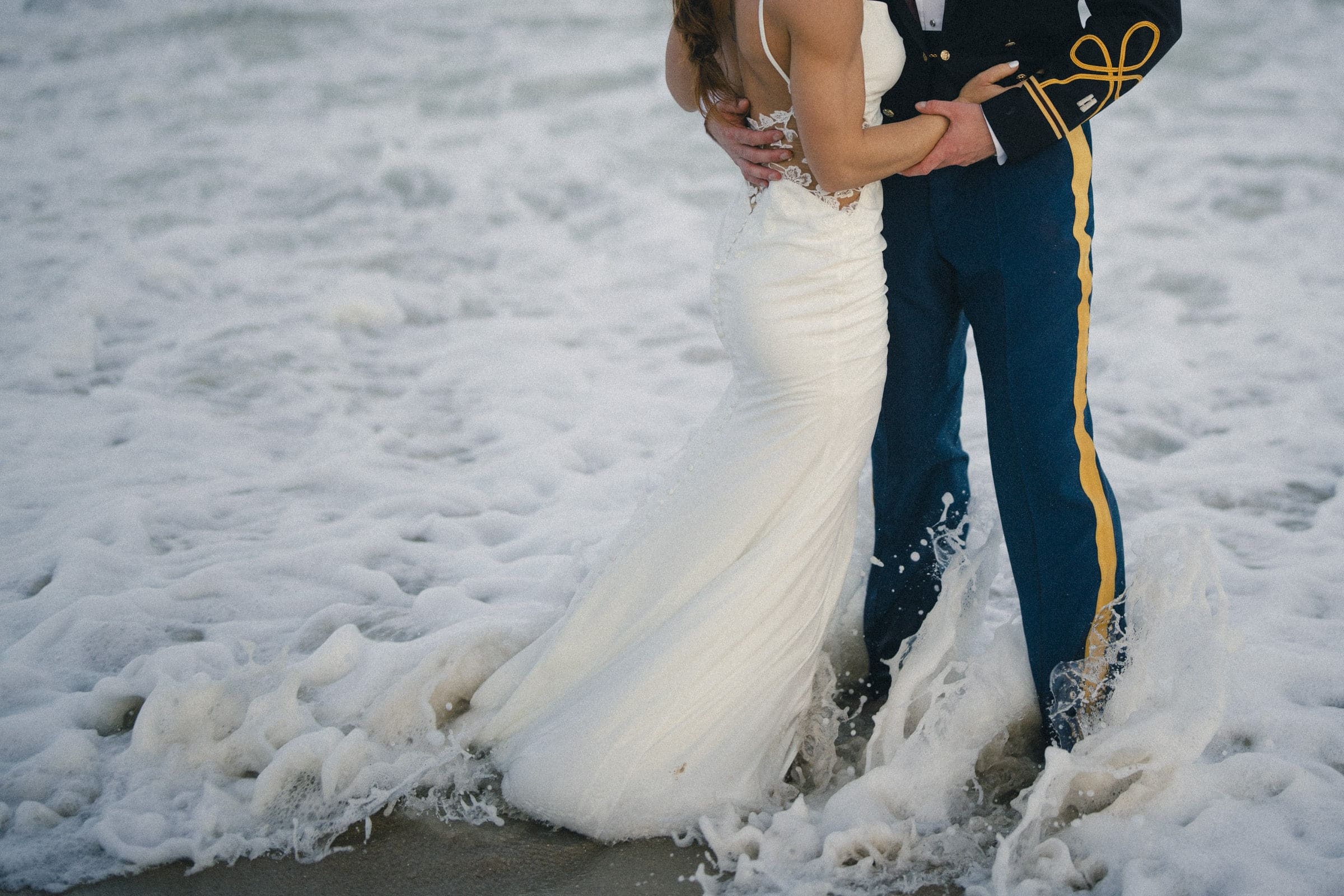 A bride in a white wedding dress and a groom in a formal military uniform stand together at the edge of the ocean, with waves washing over their feet.