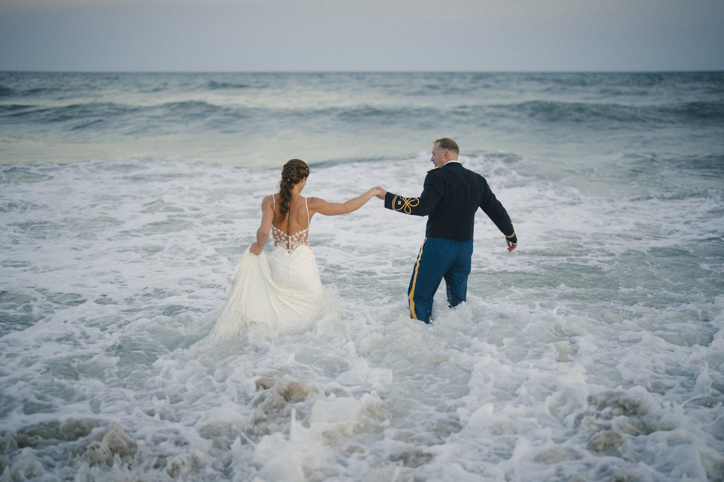 A bride and groom hold hands as they walk into the ocean waves, with the bride in a white dress and the groom in a dark military uniform, viewed from behind.