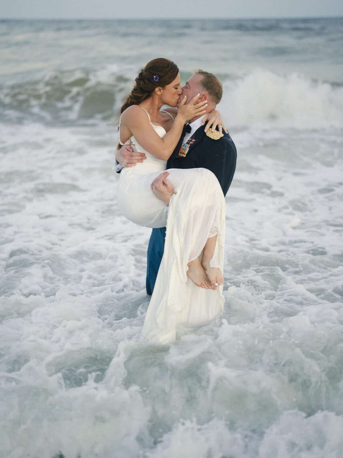 A groom in a navy suit and a bride in a white dress share a kiss as he lifts her in his arms, standing together in the surf of a beach with waves around them.