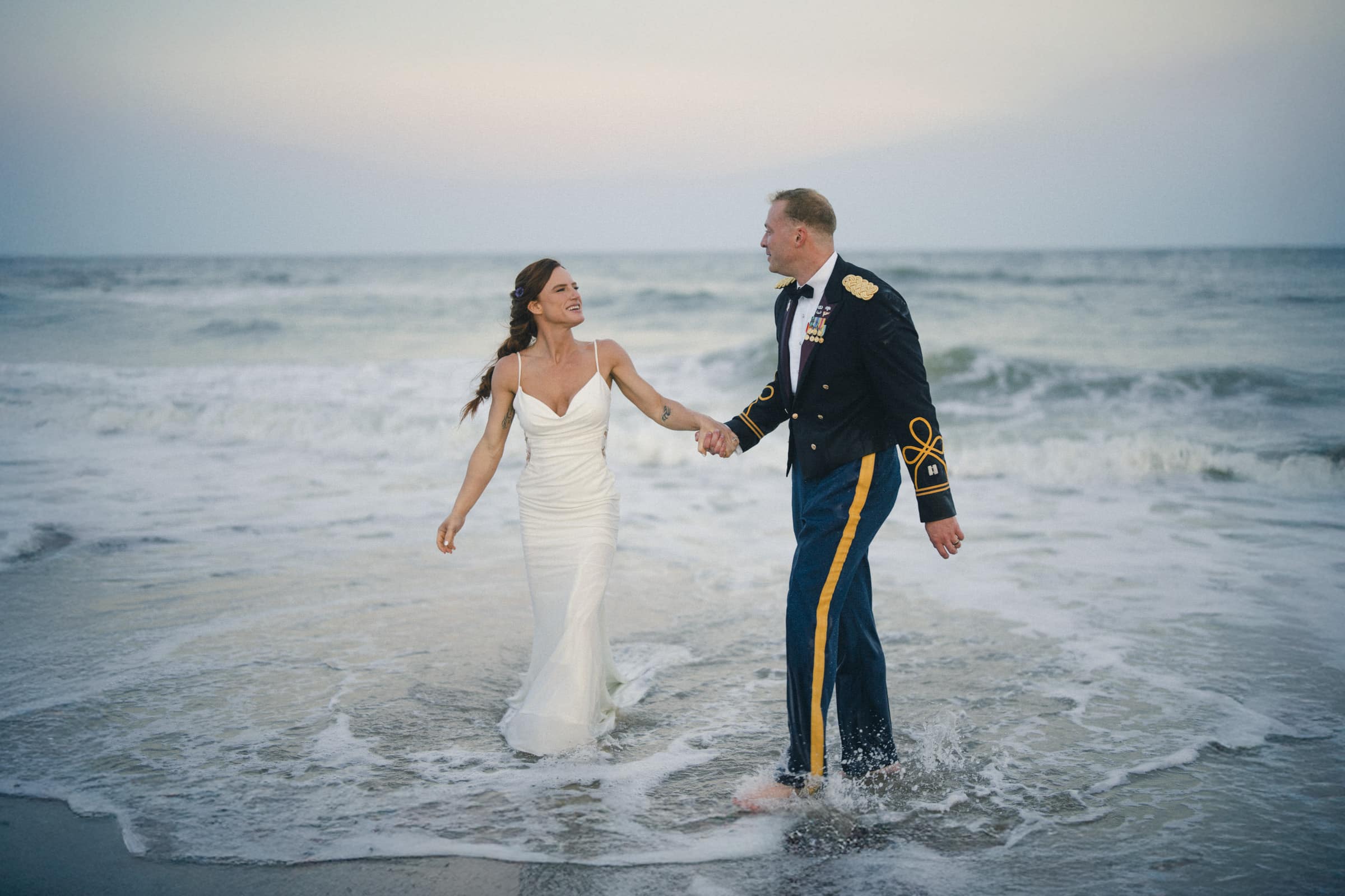 A bride in a white dress and a groom in a military uniform hold hands and smile while standing barefoot in shallow ocean waves on a beach at sunset.