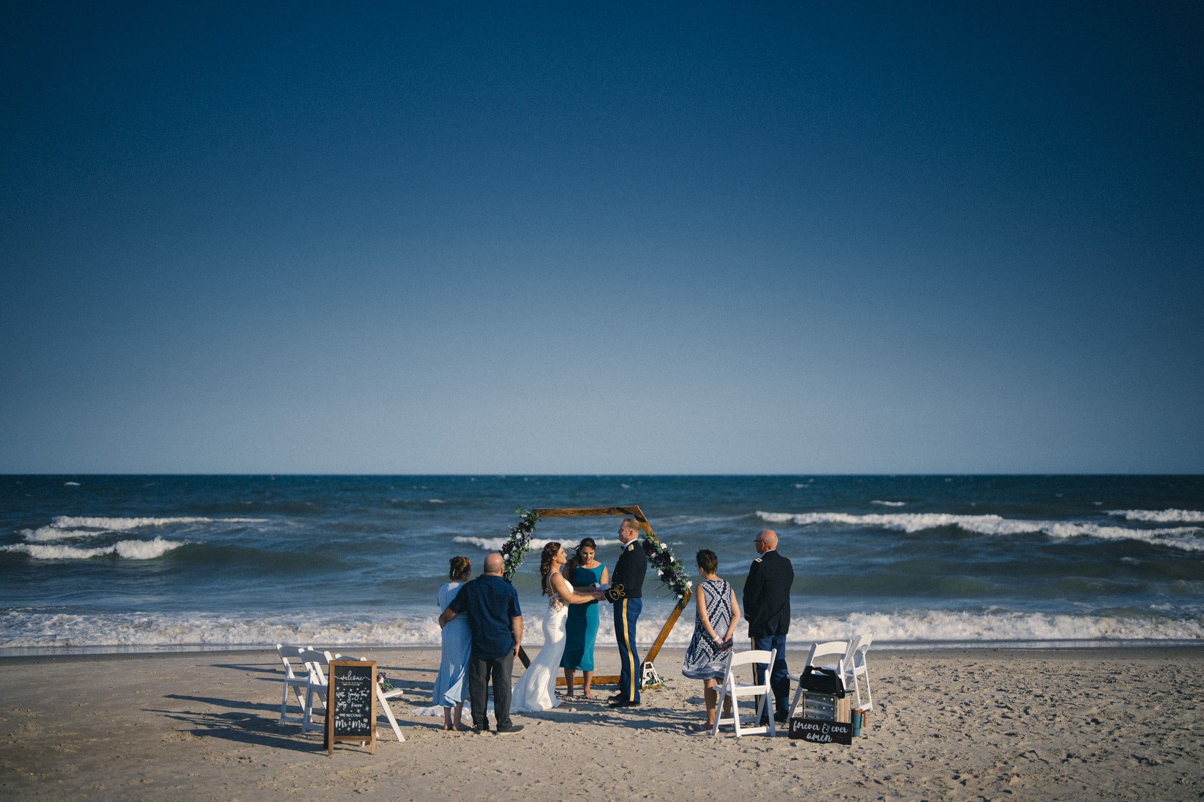 A small wedding ceremony is held on a sandy beach, with a couple standing under an arch, exchanging vows as a few guests look on. The ocean and blue sky create a peaceful backdrop.