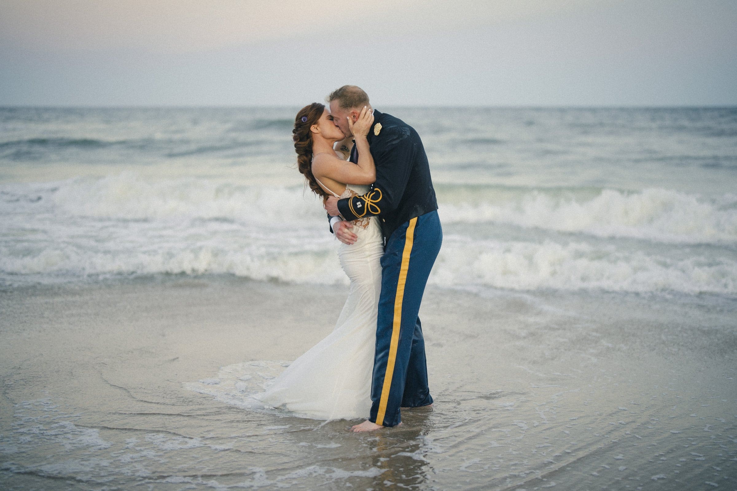 A bride in a white dress and a groom in a military uniform share a kiss while standing barefoot on the wet sand at the edge of the ocean. Waves gently roll in behind them under a soft, pale sky.