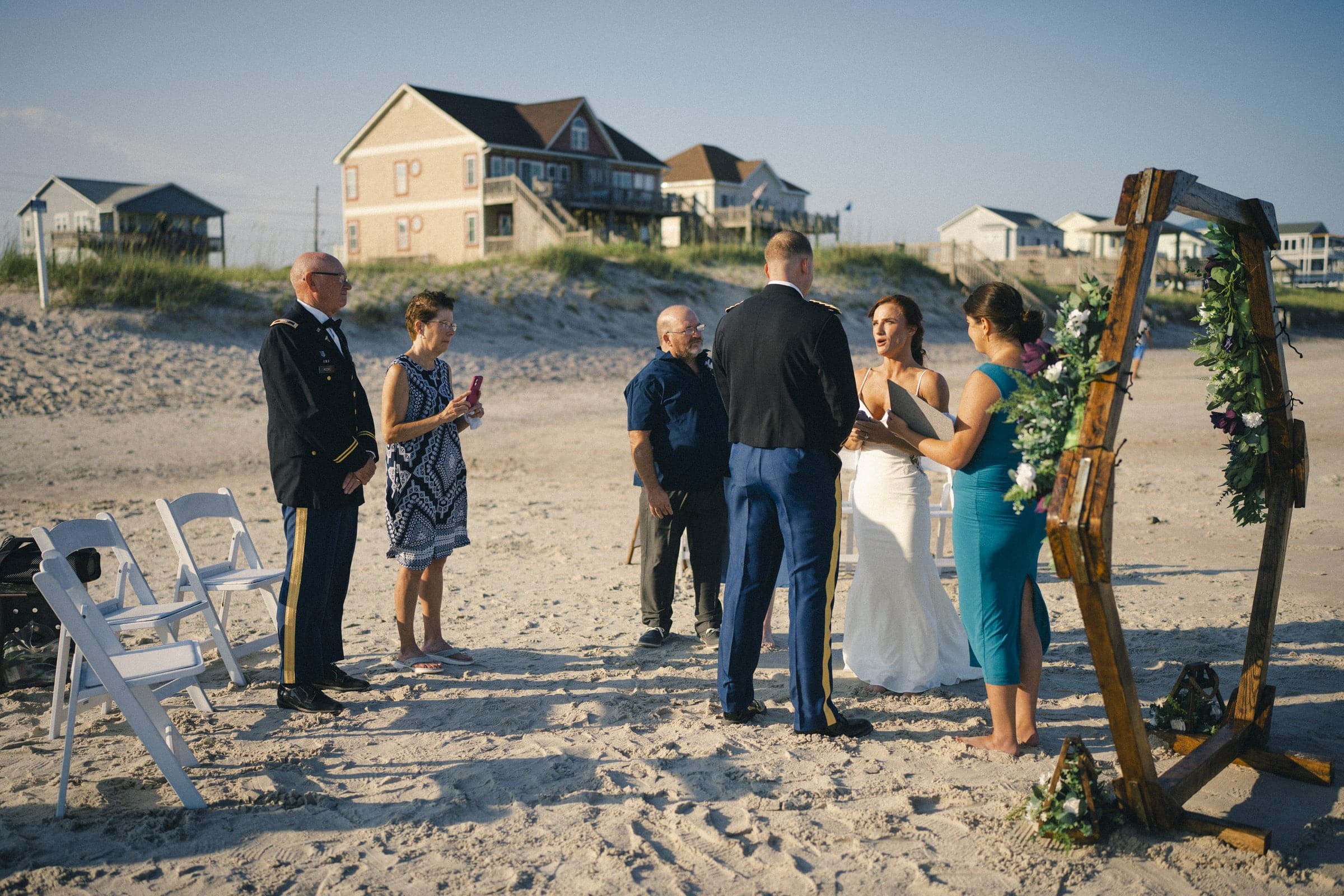A small wedding ceremony takes place on a sandy beach. The bride and groom stand under a decorated wooden arch, surrounded by a few guests, with beach houses in the background and clear blue skies above.