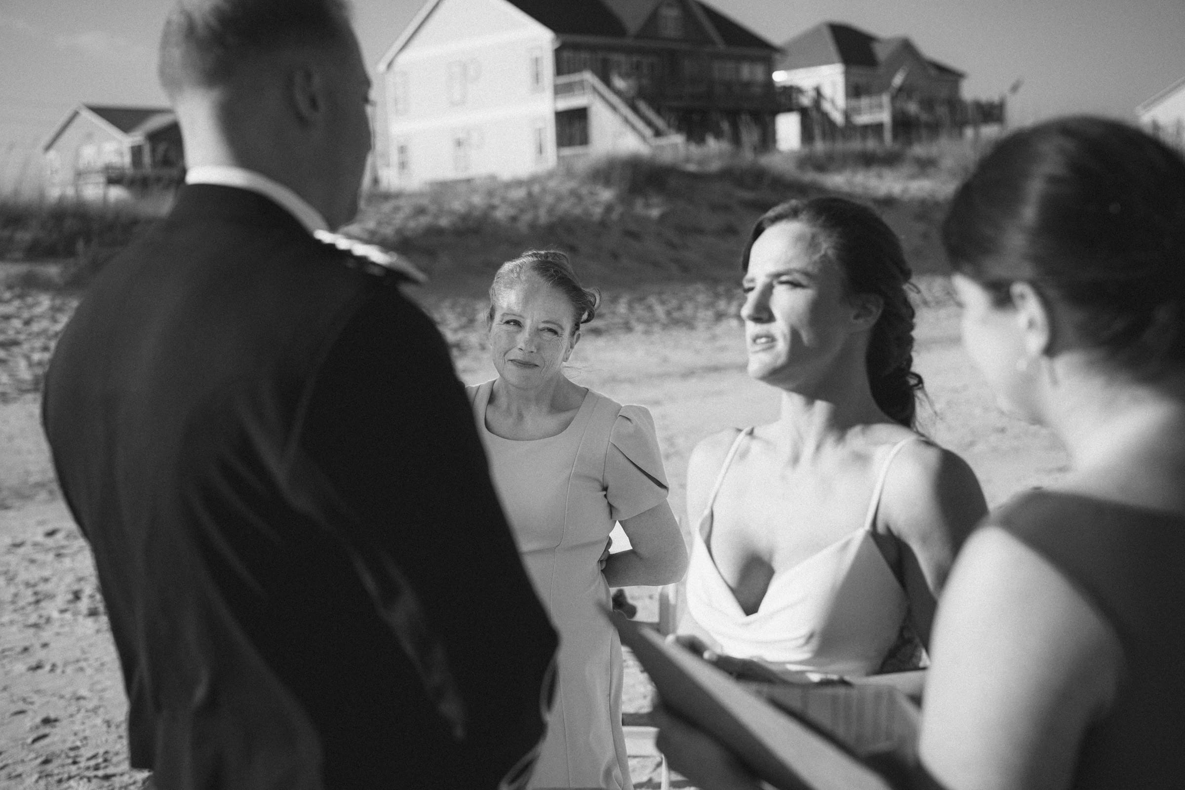 A bride and groom stand on a beach during a wedding ceremony, with two women beside them. Houses and dunes are visible in the background. The image is in black and white.
