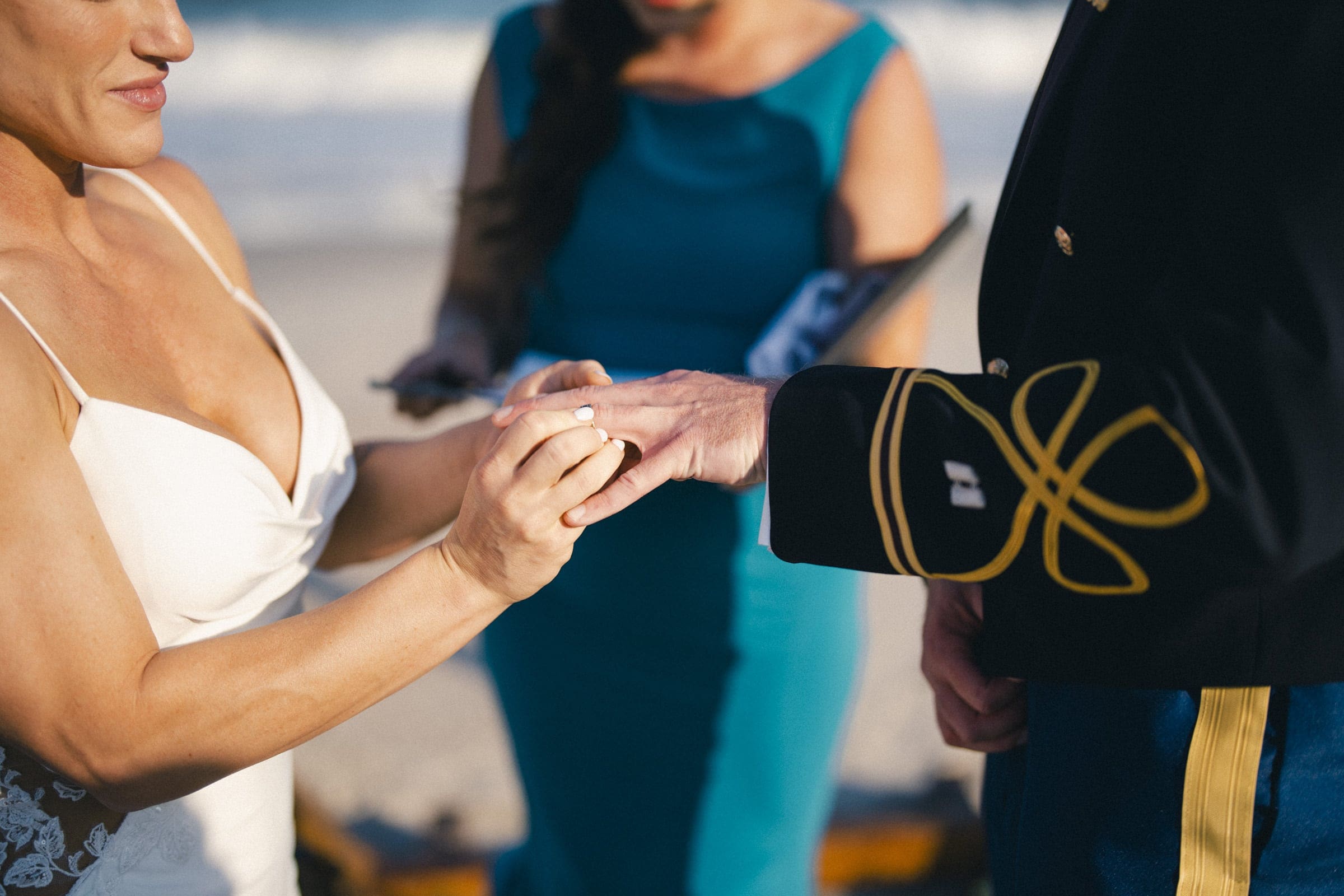 A bride places a ring on a groom’s finger during a beach wedding ceremony. The groom wears a formal military uniform and an officiant stands behind them, holding a book. The ocean is visible in the background.