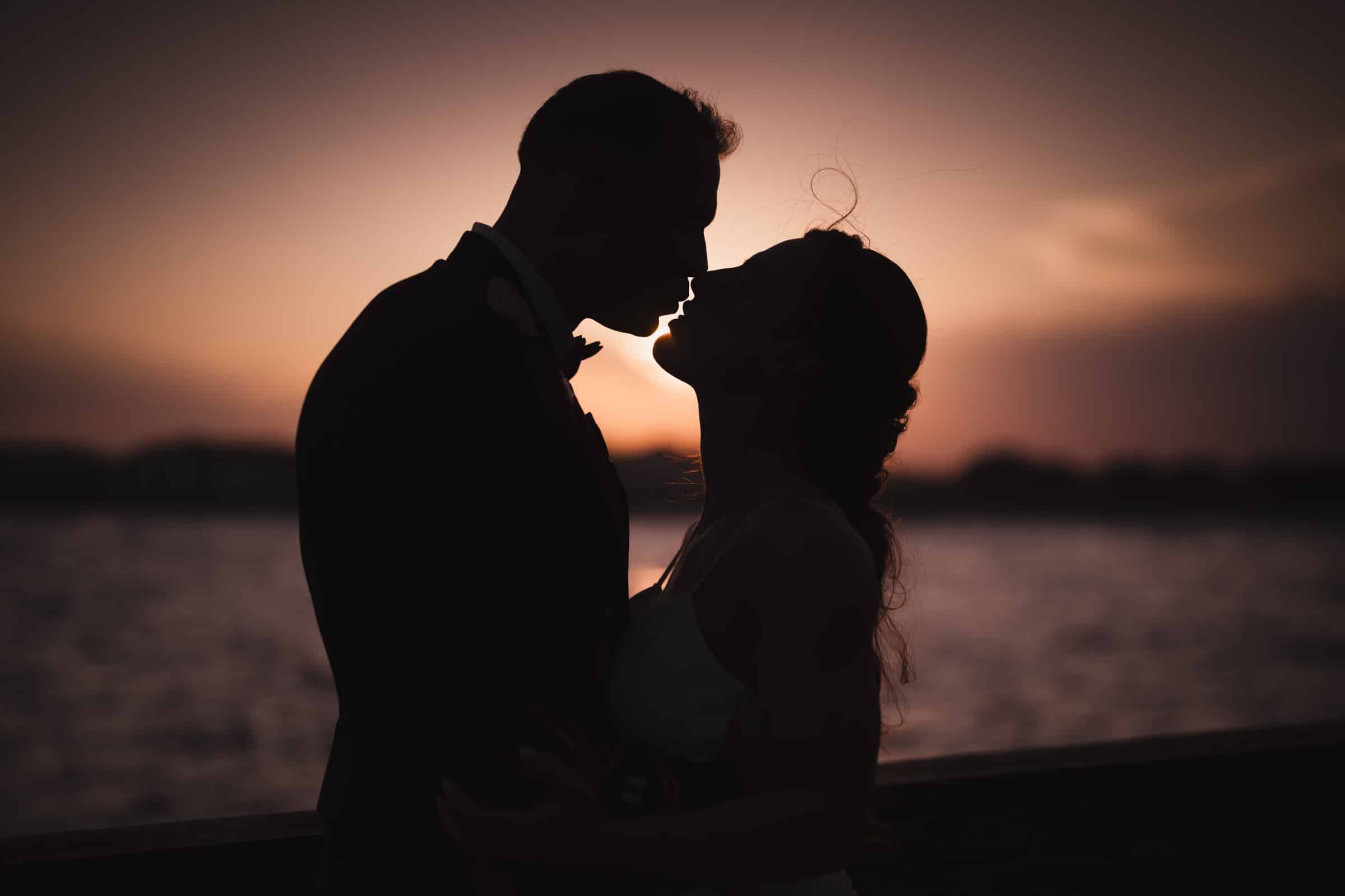 A silhouetted couple stands close together, about to kiss, against a sunset sky by the water, creating a romantic and intimate scene.