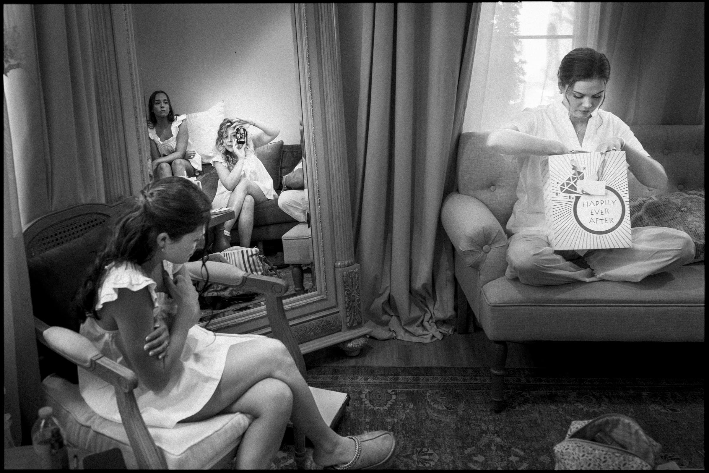 A woman opens a gift bag labeled Happily Ever After while three other women, visible in a mirror’s reflection, sit and chat in a cozy, softly-lit room with curtains and comfortable seating.