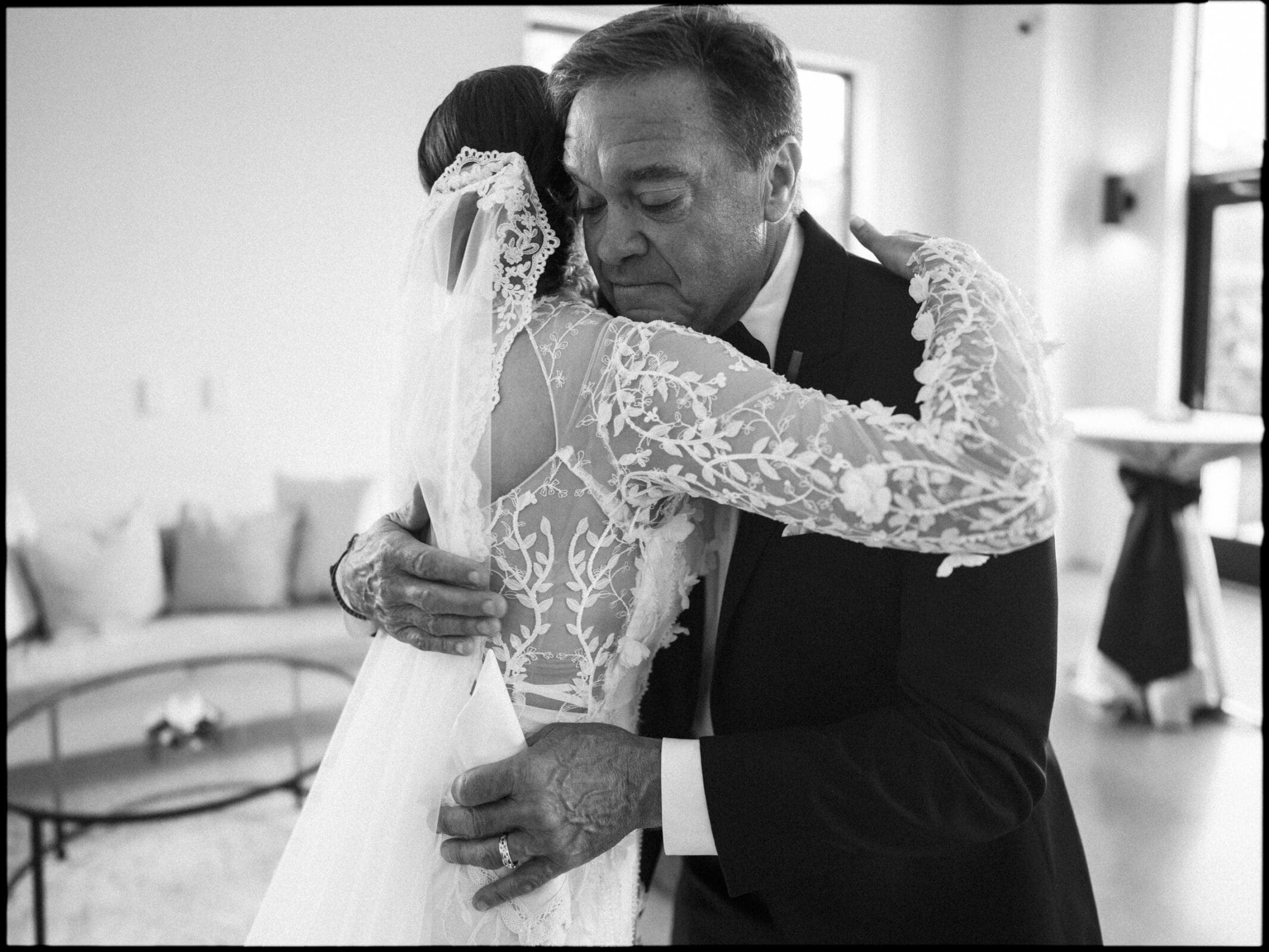A bride in a lace wedding dress embraces an older man in a suit, likely her father, in an emotional moment indoors. The scene is in black and white, capturing warmth and tenderness.