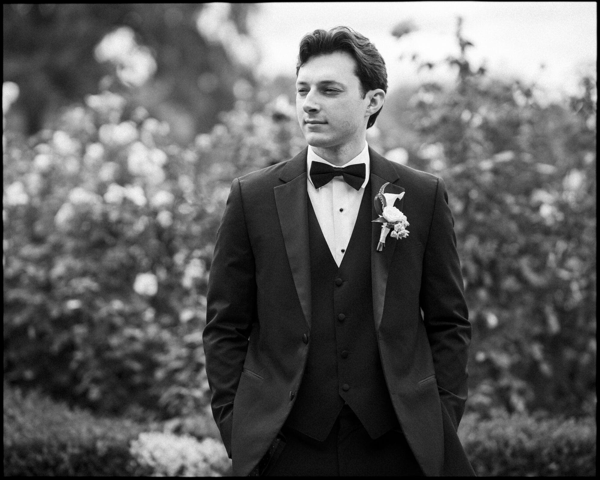 A man in a tuxedo with a bow tie and boutonniere stands outdoors, hands in pockets, looking to the side. Blurred foliage and flowers are in the background. The image is in black and white.