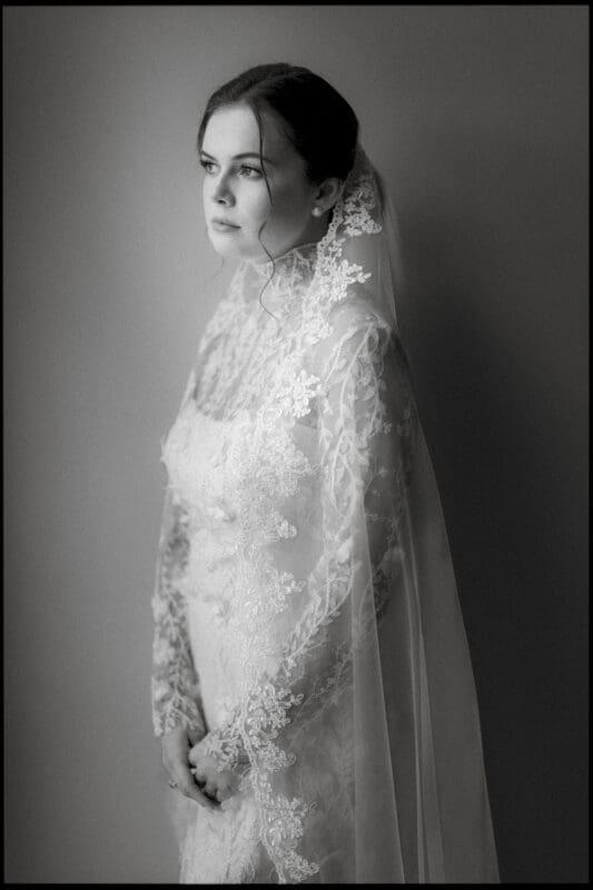 A black and white photo of a bride in a lace wedding dress and veil, standing against a plain background, gazing thoughtfully to the side with hands gently clasped.
