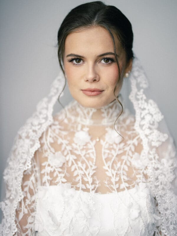 A bride with dark hair and a lace veil looks at the camera. She wears a white dress with floral lace details and has soft makeup. The background is a neutral, light color.