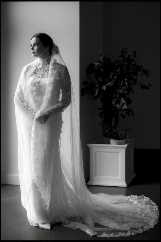 A bride in a long, lace wedding dress and veil stands by a window, looking out thoughtfully. A potted plant in a white container is seen in the background. The image is in black and white.