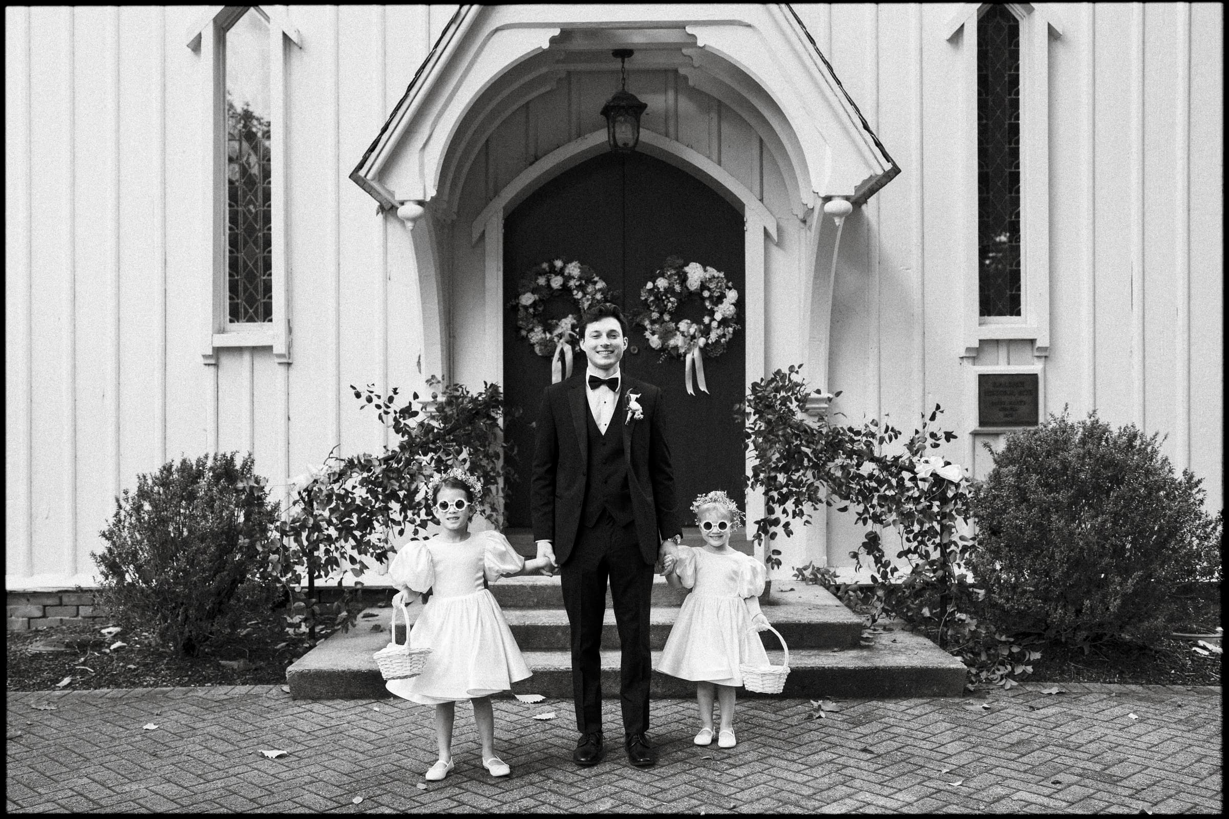 A man in a tuxedo stands smiling between two young girls in dresses and sunglasses, holding baskets, in front of a church decorated with wreaths. The photo is black and white.