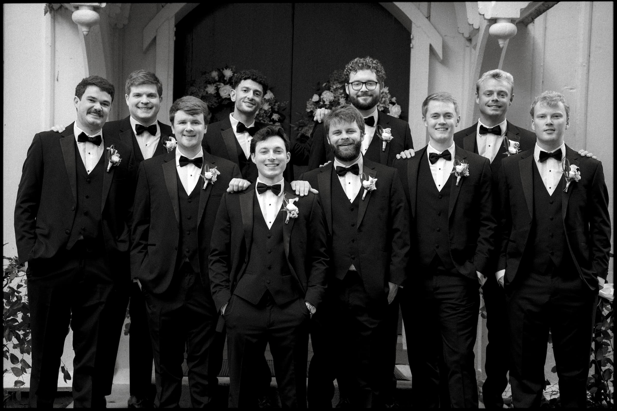 A group of eleven men dressed in black tuxedos and bow ties pose together, smiling, in front of a decorated archway with flowers. It appears to be a formal event, such as a wedding. The photo is in black and white.