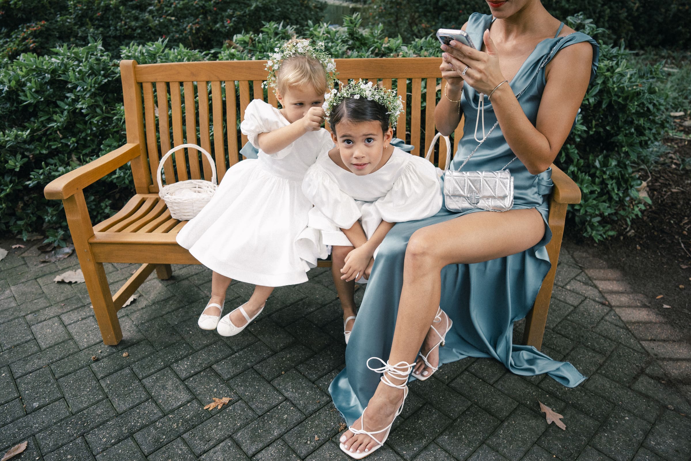 Two young girls in white dresses and flower crowns sit on a wooden bench, one touching the others hair. A woman in a teal dress sits beside them, looking at her phone. All three have baskets, likely at a formal event.