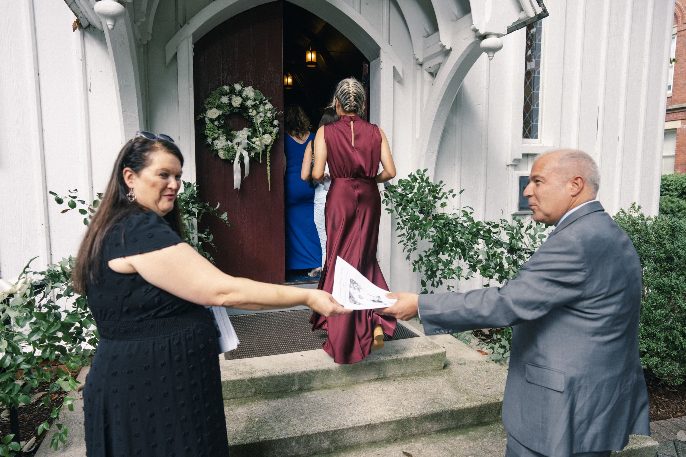 A woman in a black dress and a man in a gray suit hand out programs at the entrance of a church, as guests walk inside through a doorway decorated with a white floral wreath.