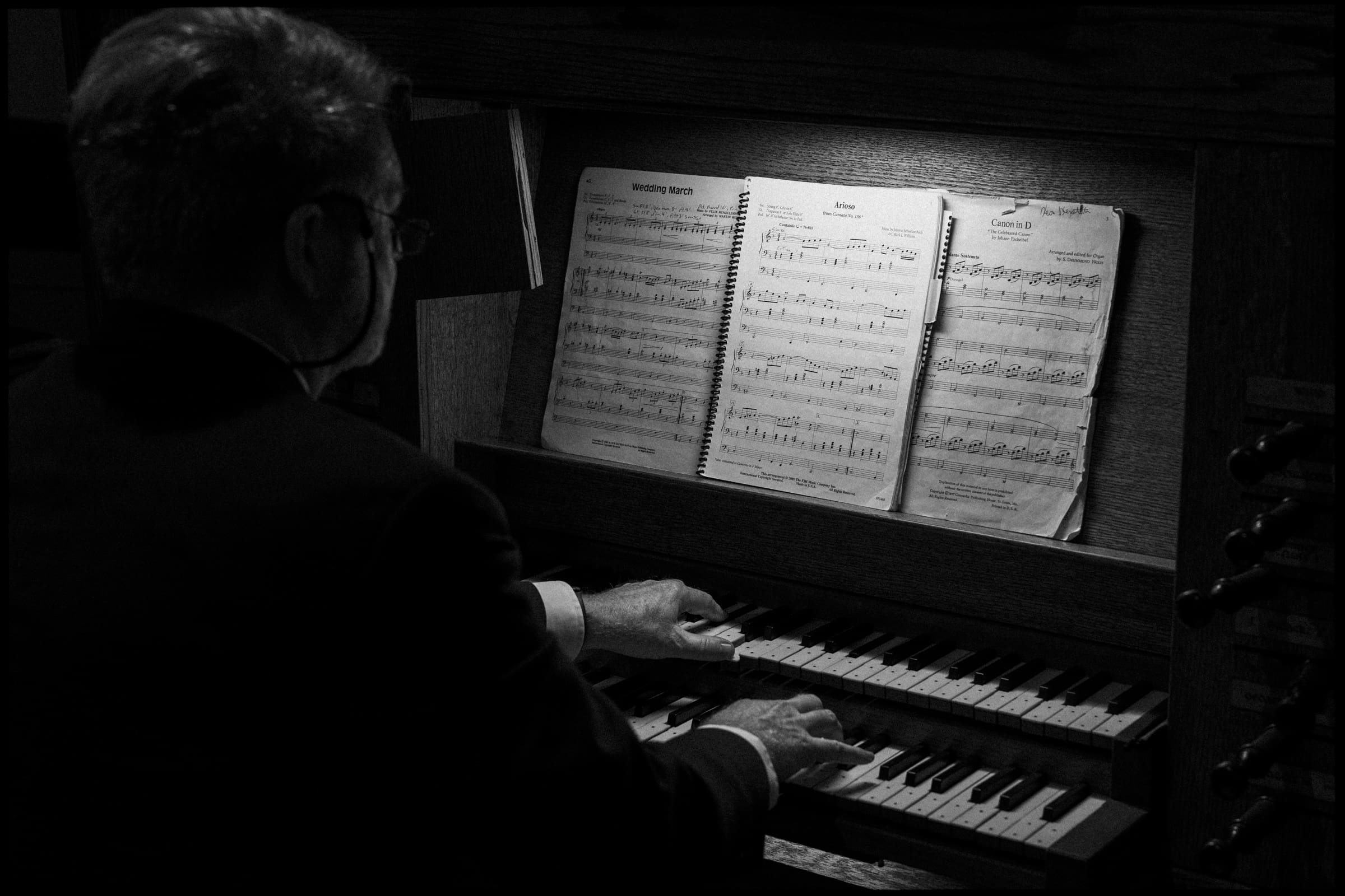 A person in a suit is playing a pipe organ, reading sheet music illuminated by a light. The photo is in black and white, highlighting the hands on the keys and the open music books.