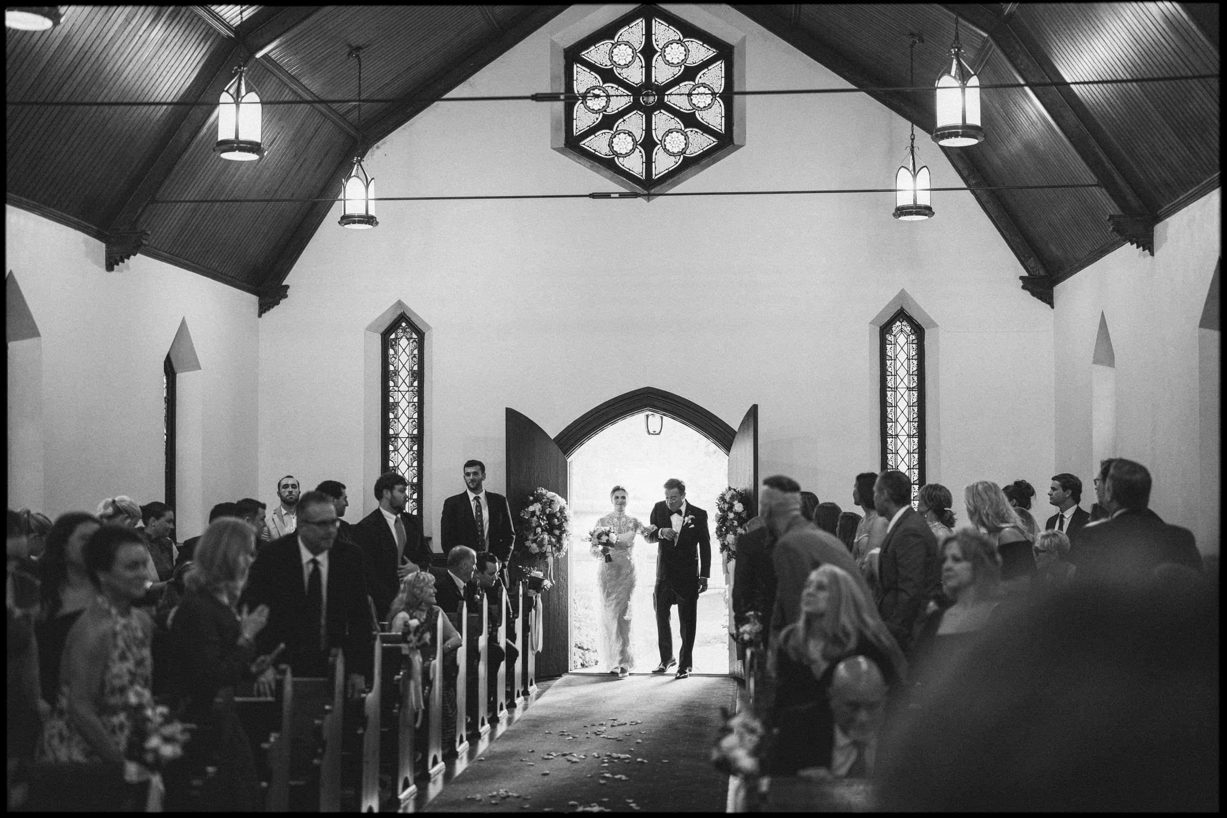 A black and white photo of a bride walking down the aisle of a church with her escort, surrounded by seated and standing guests, under arched ceilings and stained glass windows.