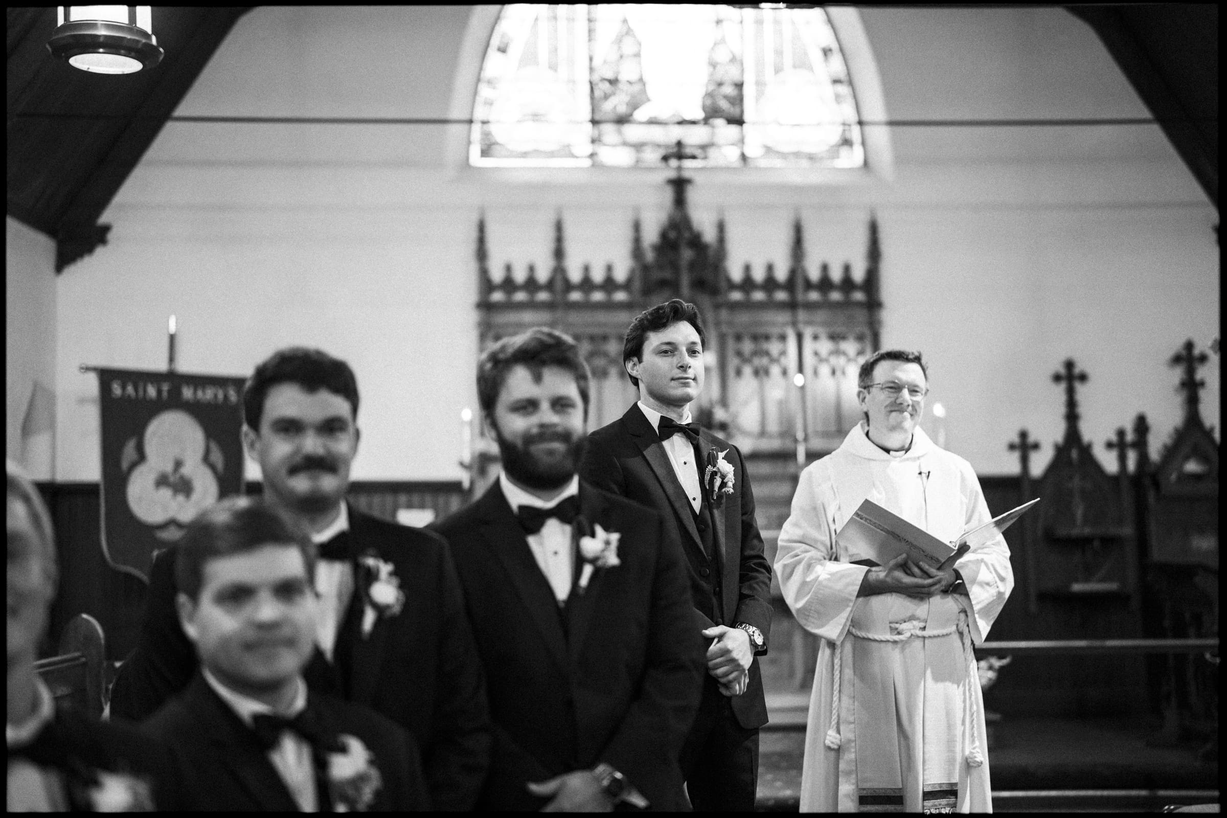 A groom in a tuxedo stands with groomsmen and a smiling officiant at the altar of a church, with stained glass windows and wooden pews in the background. The scene is black and white.