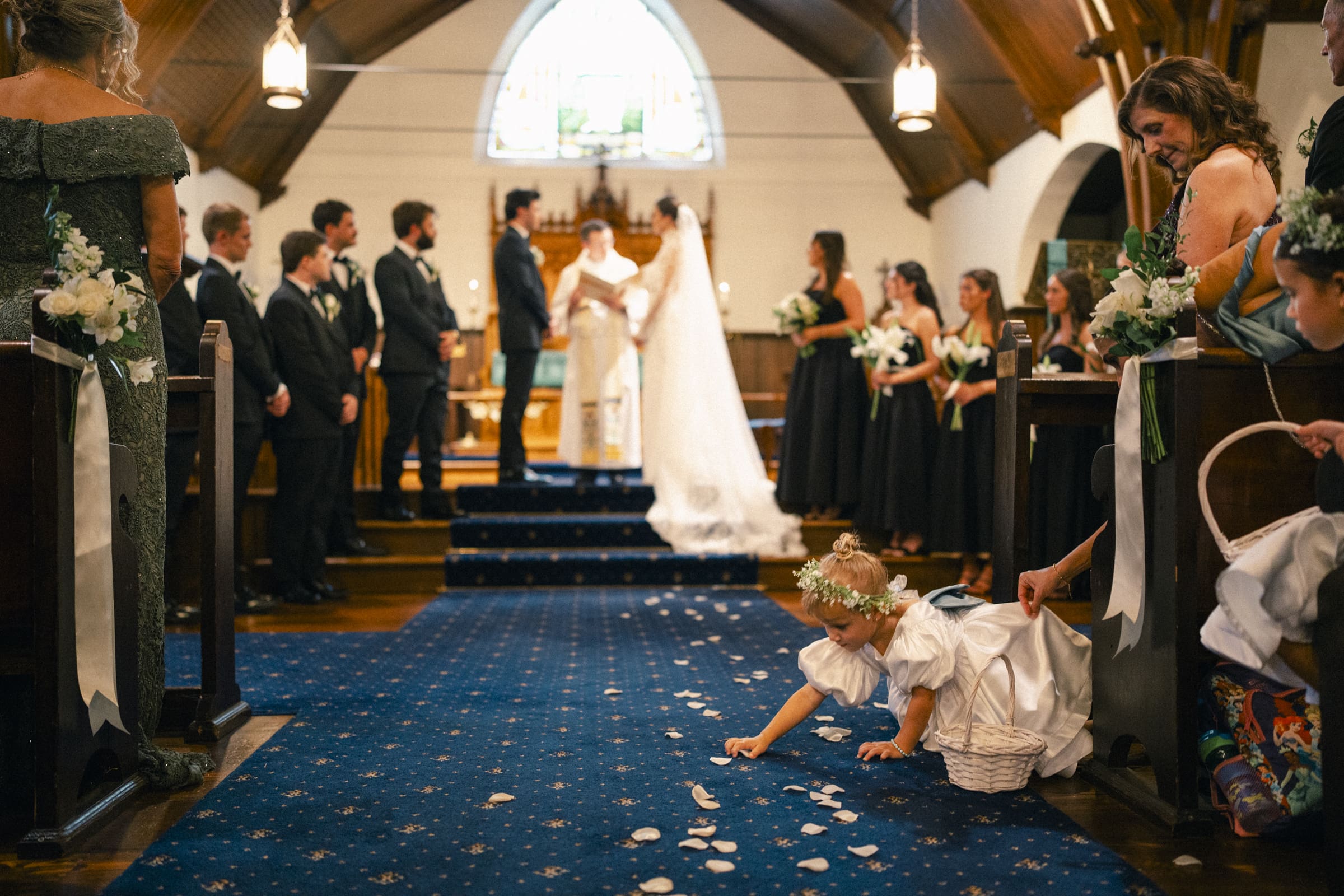 A young flower girl in a white dress picks up petals on the aisle of a church as a wedding ceremony takes place at the altar, with the bride, groom, officiant, and wedding party in the background.