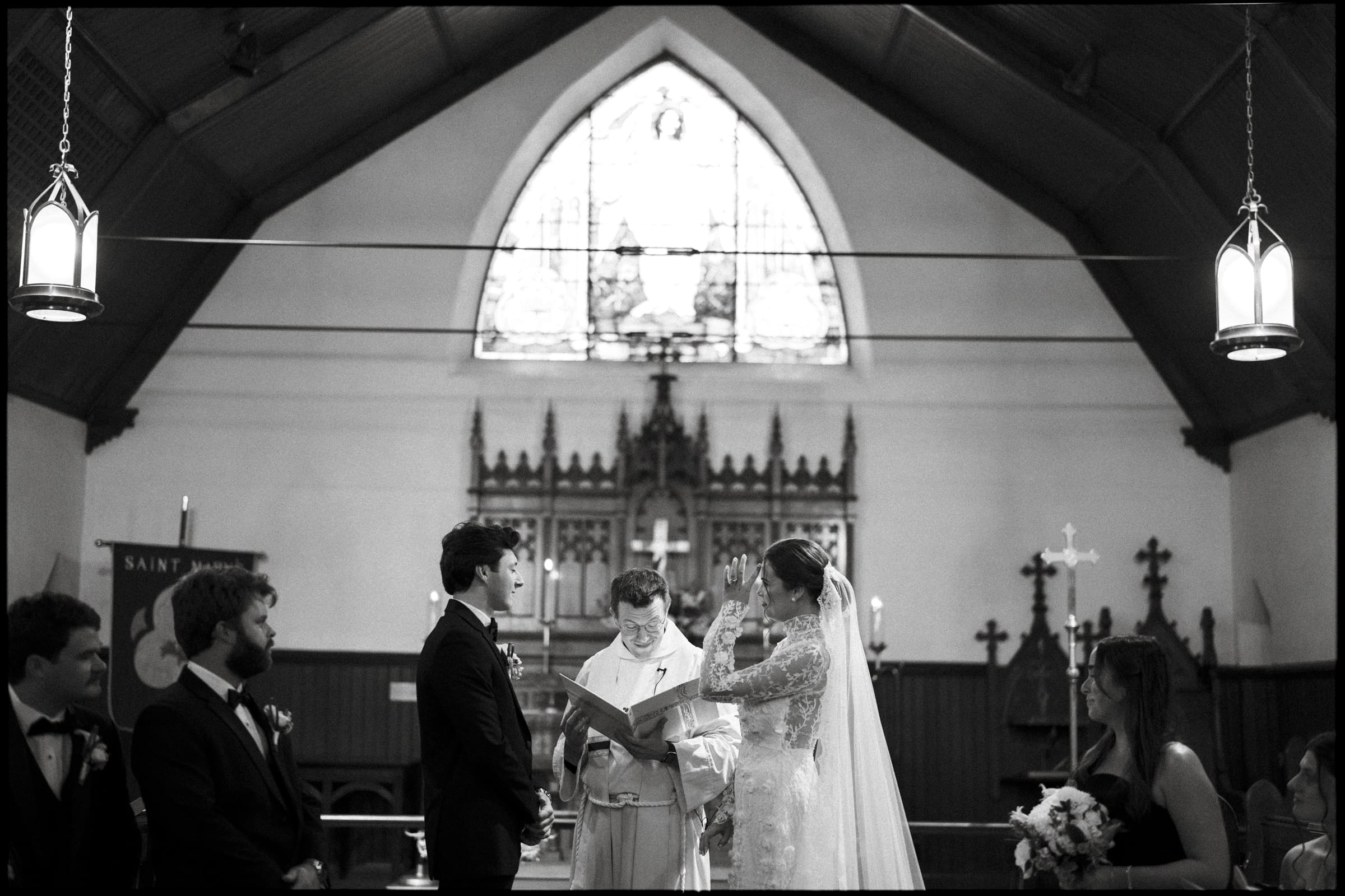 A black-and-white photo of a wedding ceremony inside a church. The bride and groom stand facing each other at the altar, with the officiant between them, and attendants on either side. Stained glass is visible in the background.