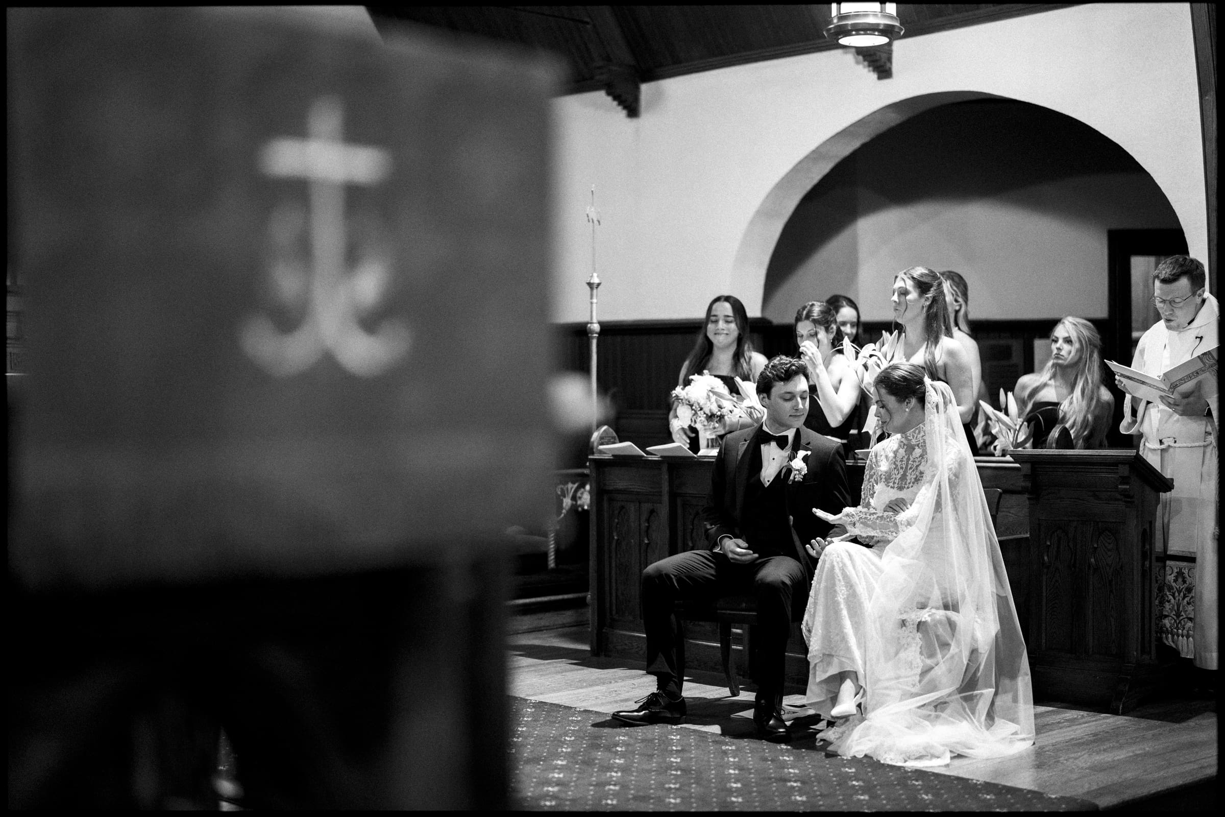 A bride and groom sit together at the altar during a church wedding ceremony, surrounded by bridesmaids and a priest, with a blurred religious symbol in the foreground. The scene is in black and white.
