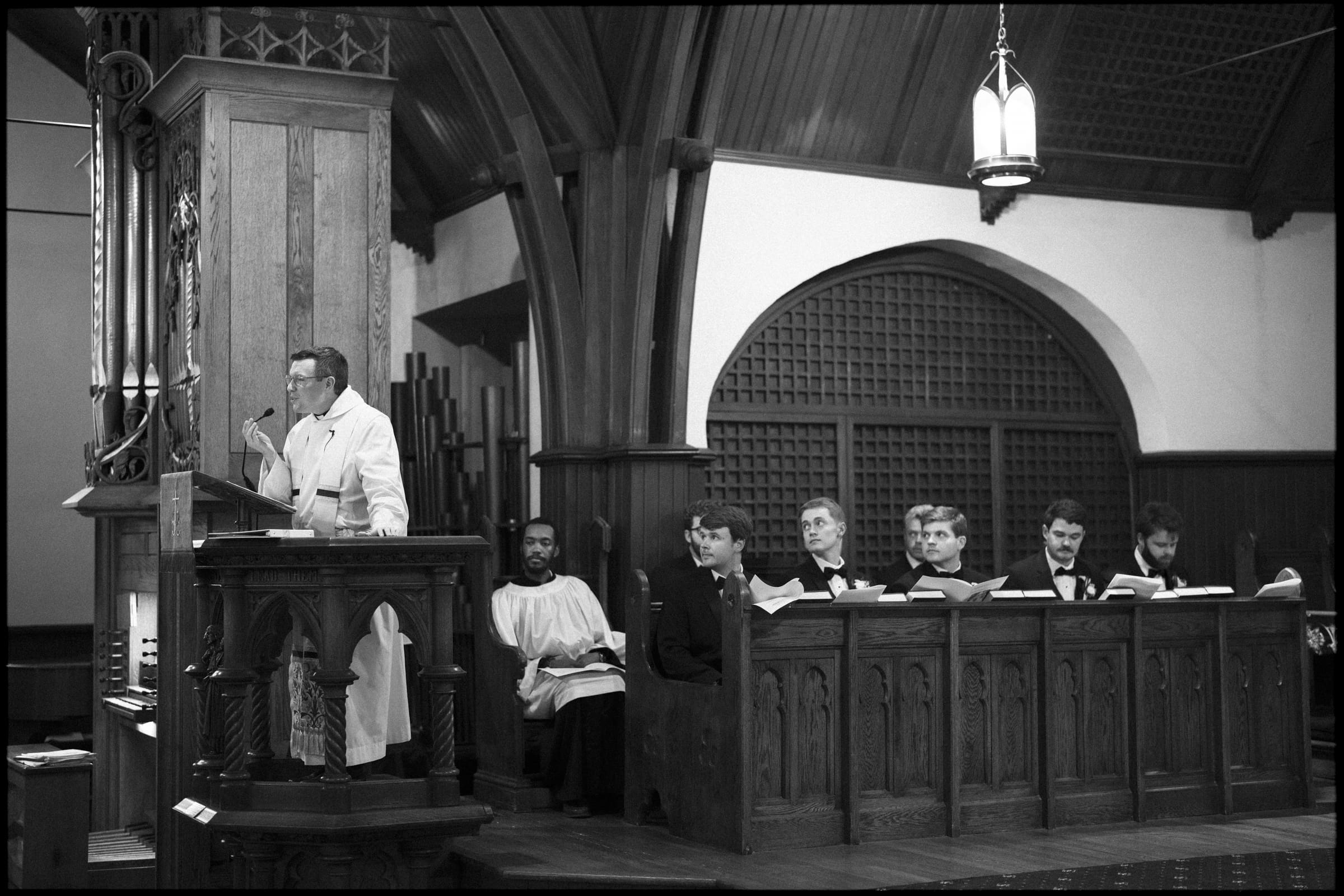 A man in a white robe speaks at a wooden church pulpit, while several people dressed in robes and suits sit in a choir box behind him, inside a church with arched ceilings and wooden panels.