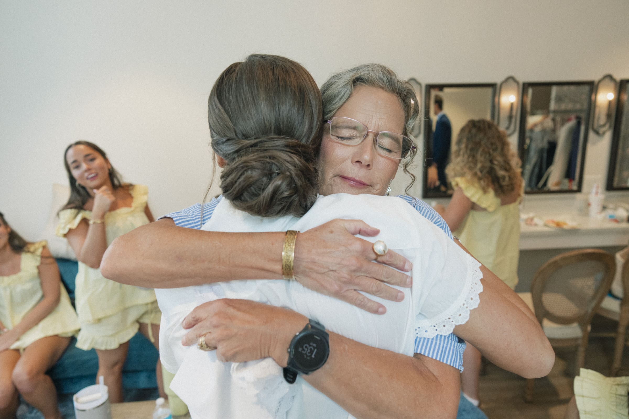 A woman with glasses closes her eyes as she hugs another woman with a low bun. In the background, several women in yellow dresses and a dressing room mirror are visible. The scene feels warm and emotional.