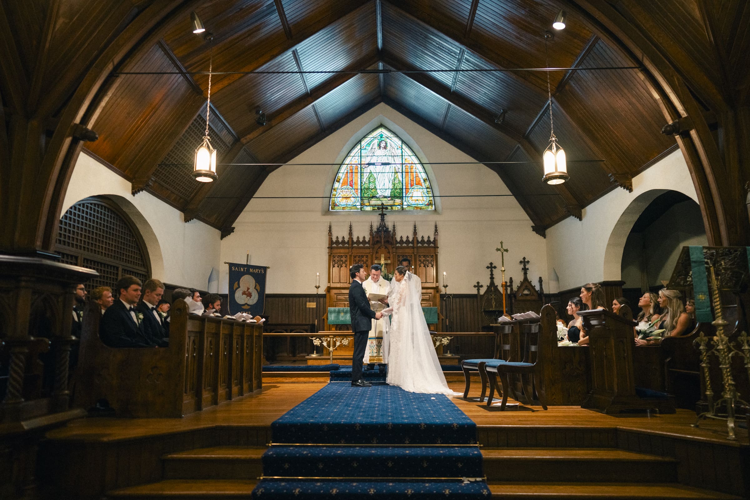 A bride and groom stand at the altar of a church with stained glass windows, exchanging vows during a wedding ceremony, surrounded by the bridal party and guests seated on either side.