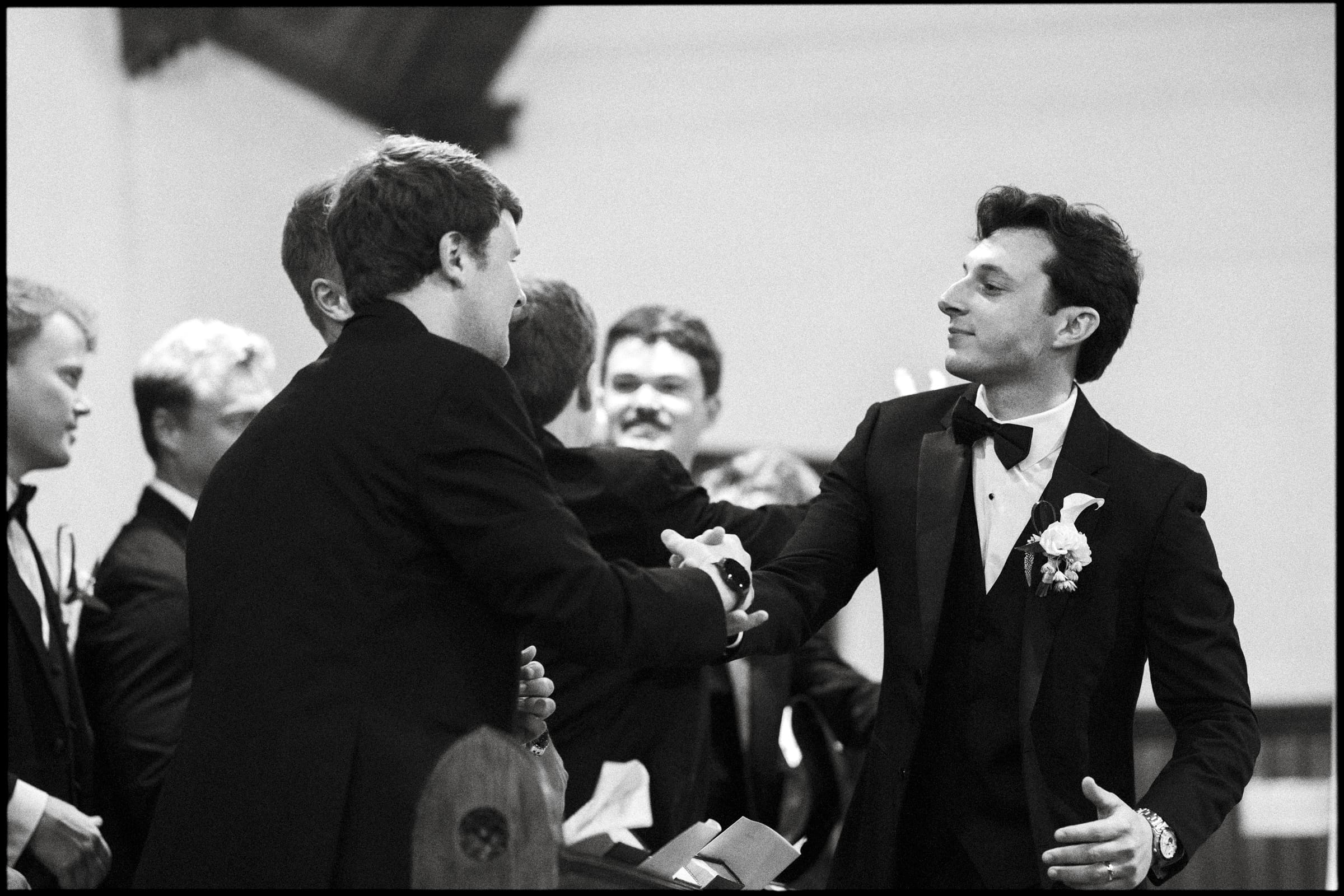 A group of men in tuxedos, one wearing a boutonnière, shake hands and smile at each other in a formal indoor setting. The atmosphere appears celebratory and supportive.