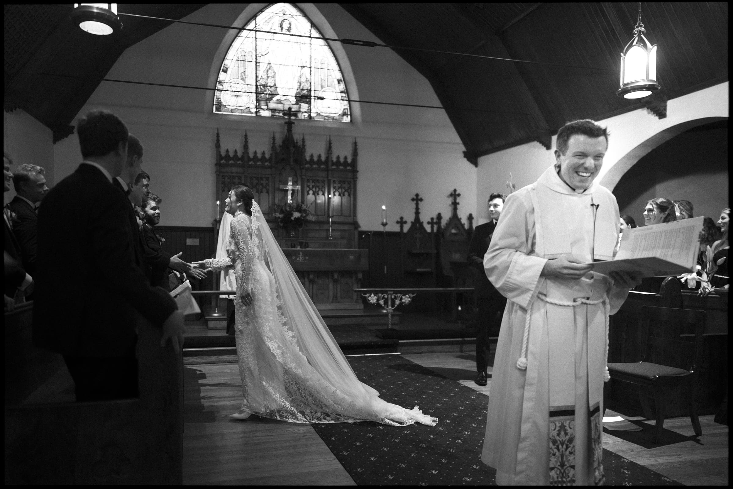 A bride in a long lace gown and veil walks down the aisle of a church, holding hands with a man. A smiling priest stands in the foreground, holding a book. Wedding guests watch from both sides.