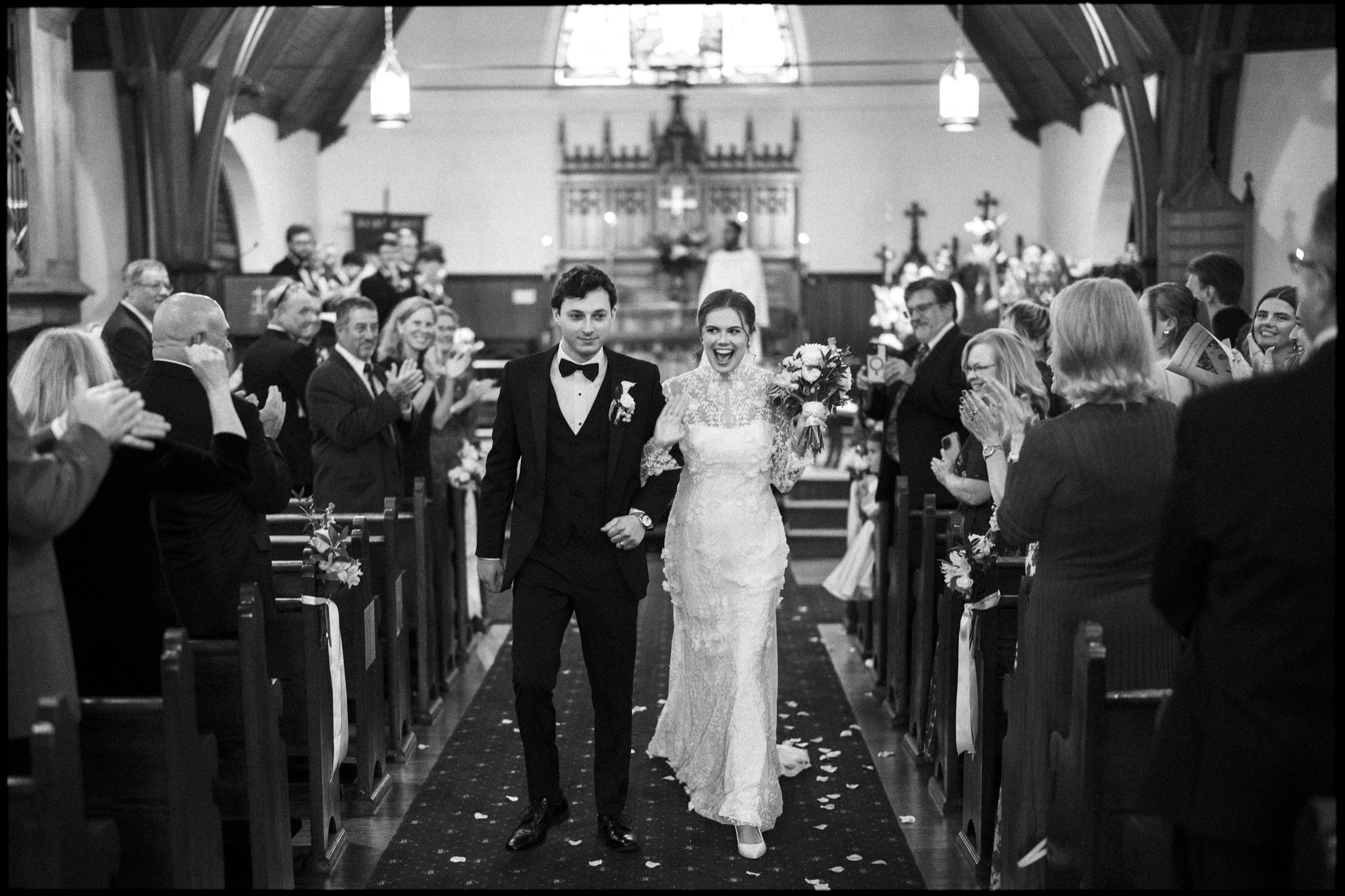 A bride and groom walk arm in arm down the aisle of a church, smiling joyfully as guests on both sides stand, applaud, and celebrate their wedding. The atmosphere is festive and filled with happiness.