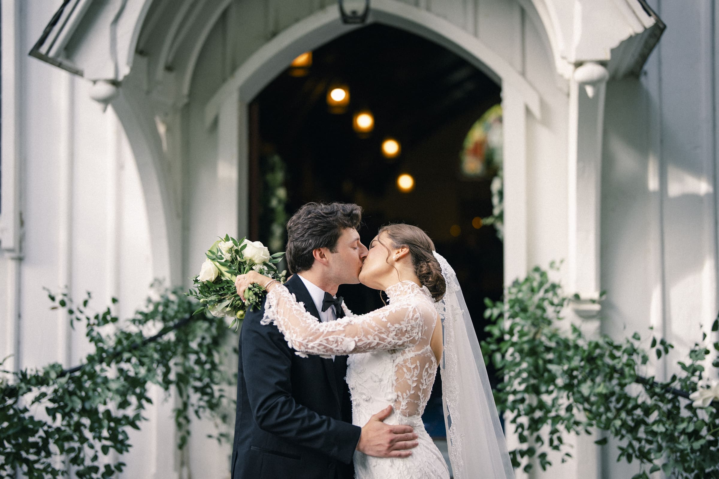 A bride and groom kiss outside a church decorated with greenery. The bride wears a white dress and veil, holding a bouquet, while the groom is in a black suit. Warm lights glow inside the church entrance behind them.
