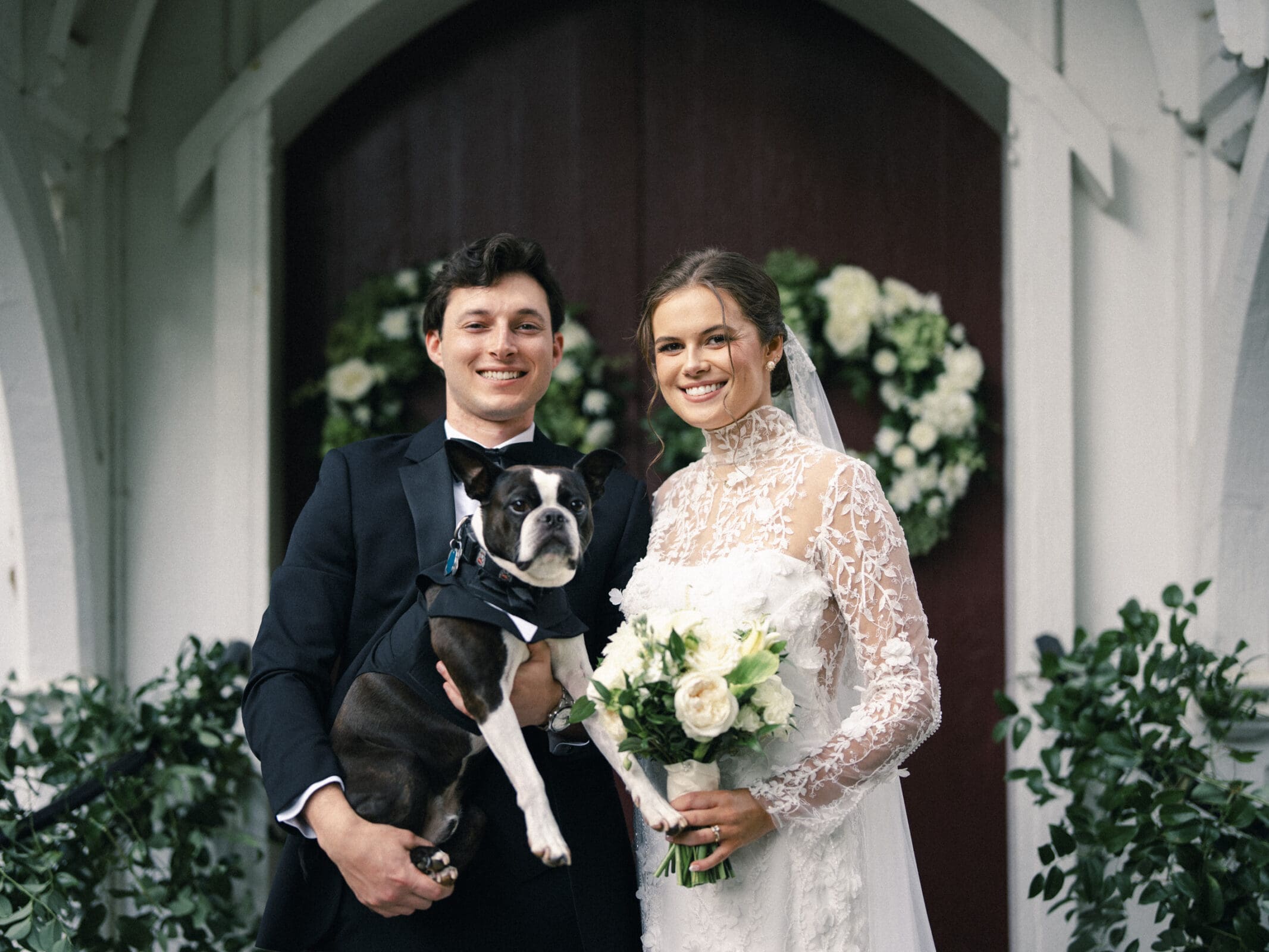 A bride and groom, both smiling, stand outside a building with floral wreaths on the doors. The groom holds a black and white dog, and the bride holds a bouquet of white flowers.