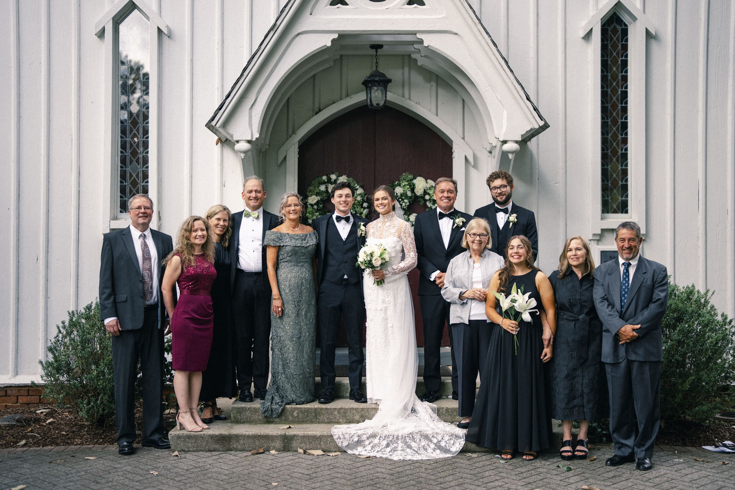A wedding party poses on church steps. The bride and groom stand in the center, surrounded by family and friends dressed in formal attire, all smiling at the camera. Greenery and white floral wreaths decorate the church door behind them.