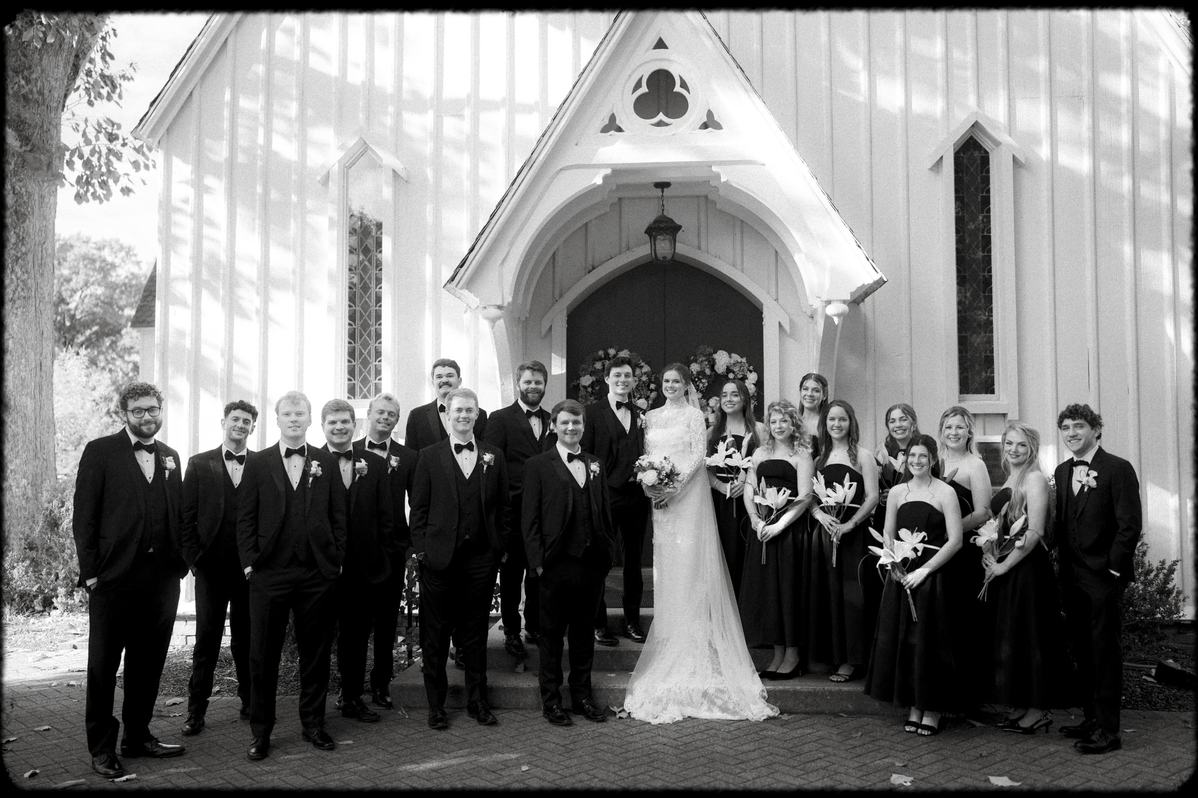 A black and white photo of a wedding party standing outside a small church. The bride and groom are in the center, surrounded by bridesmaids in dark dresses and groomsmen in tuxedos, all smiling at the camera.